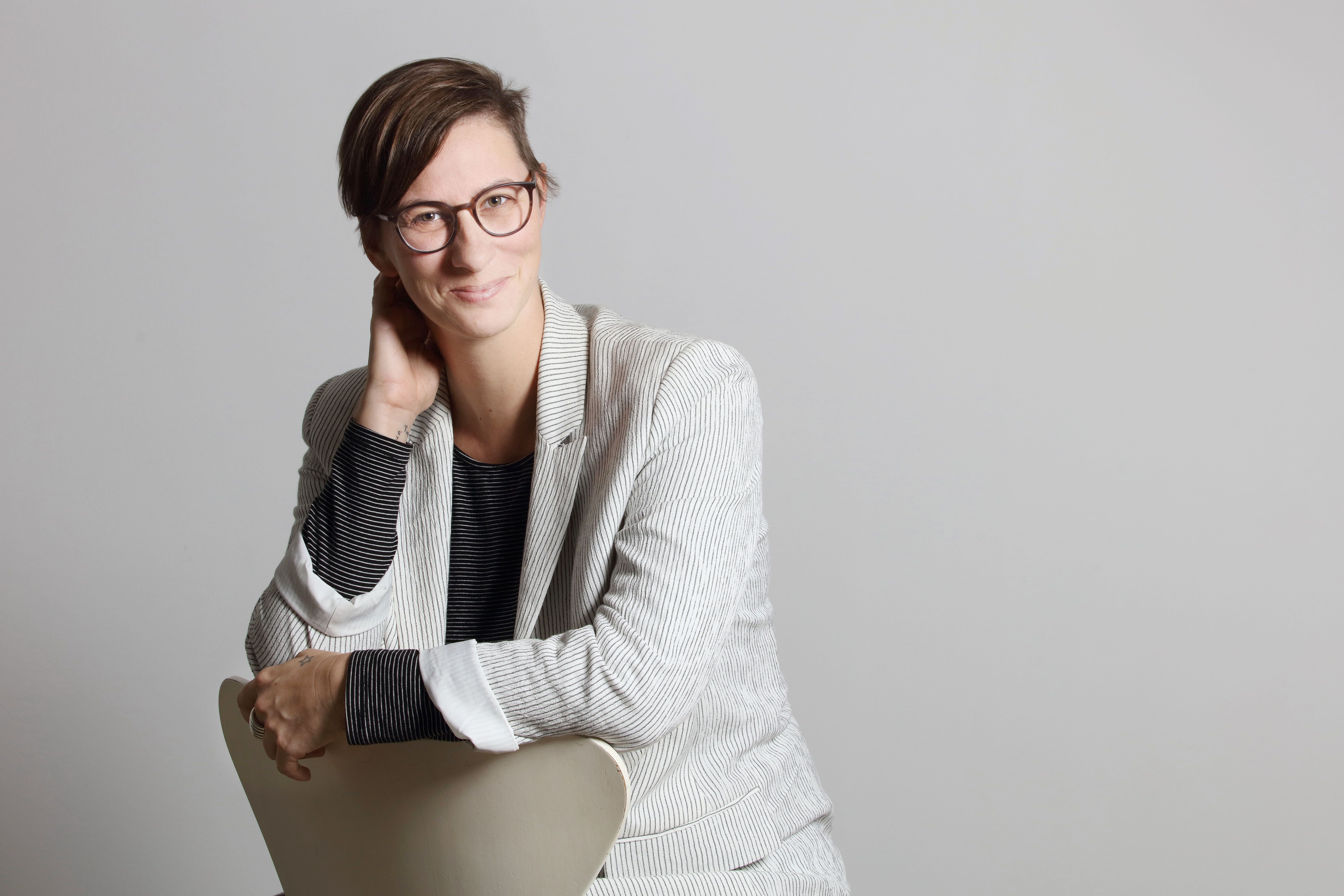Person mit Brille und hellem Blazer sitzt lächelnd auf einem Stuhl vor grauem Hintergrund.
