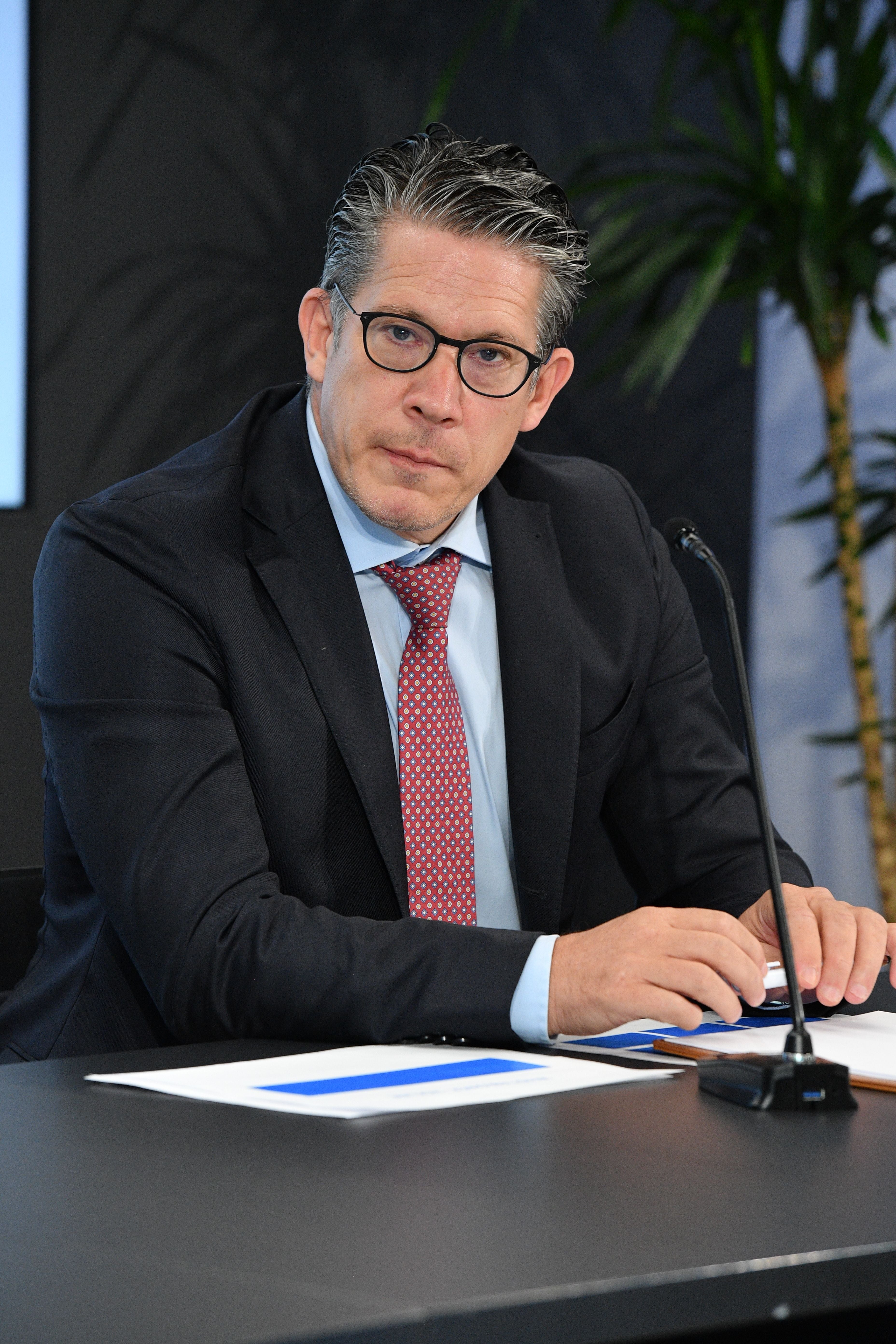 Mann im Anzug mit Brille sitzt bei einer Pressekonferenz am Tisch mit Mikrofon und Dokumenten.