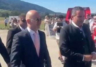 Mehrere Männer im Anzug gehen bei sonnigem Wetter im Freien, Menschen und Berge im Hintergrund.