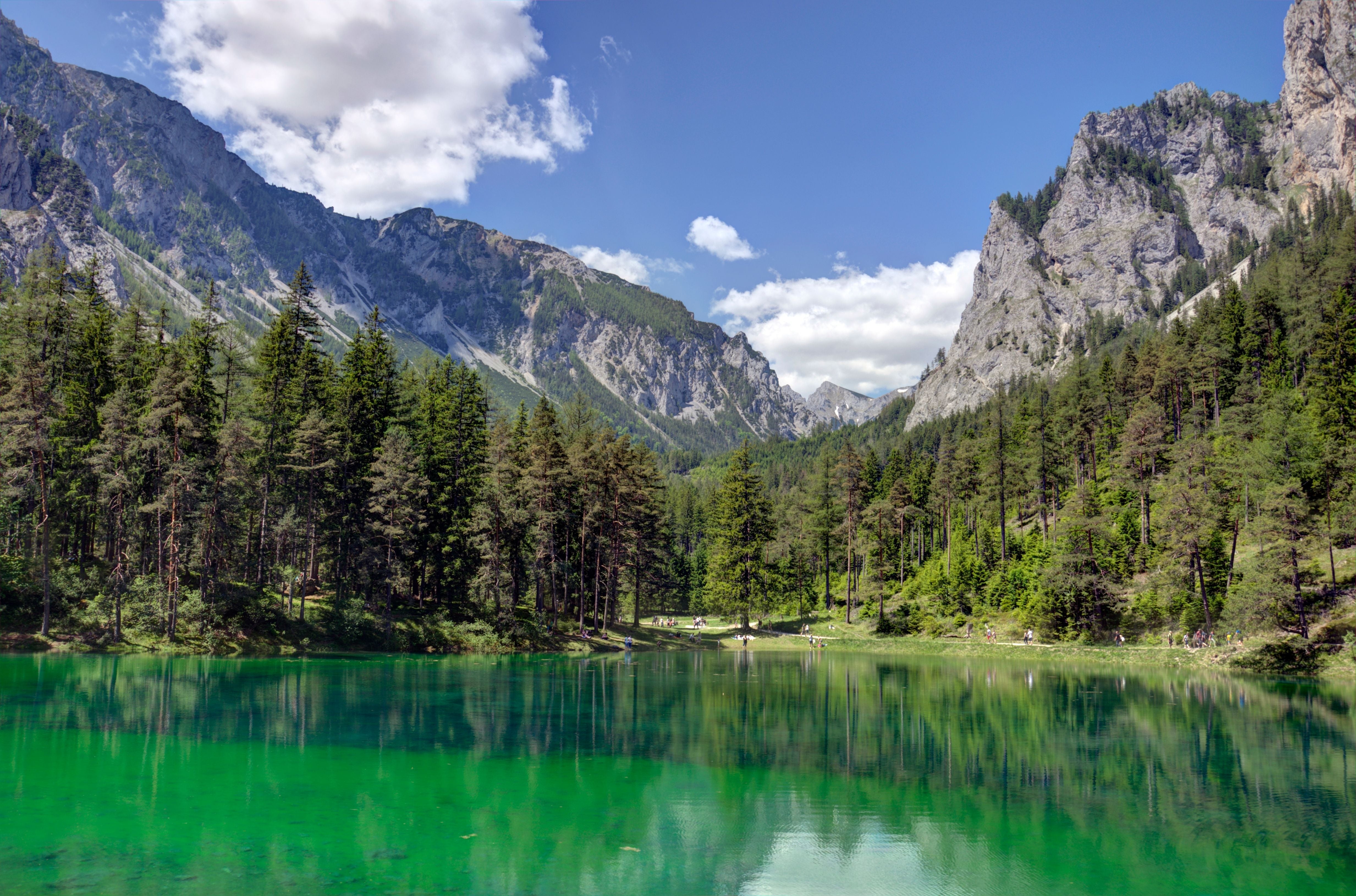 Grüner See mit smaragdgrünem Wasser, umgeben von Wäldern und hohen Bergen bei blauem Himmel.