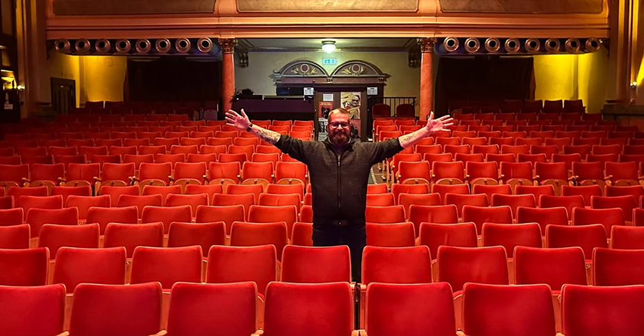 Mann steht mit ausgebreiteten Armen in einem leeren, rot gepolsterten Theatersaal.