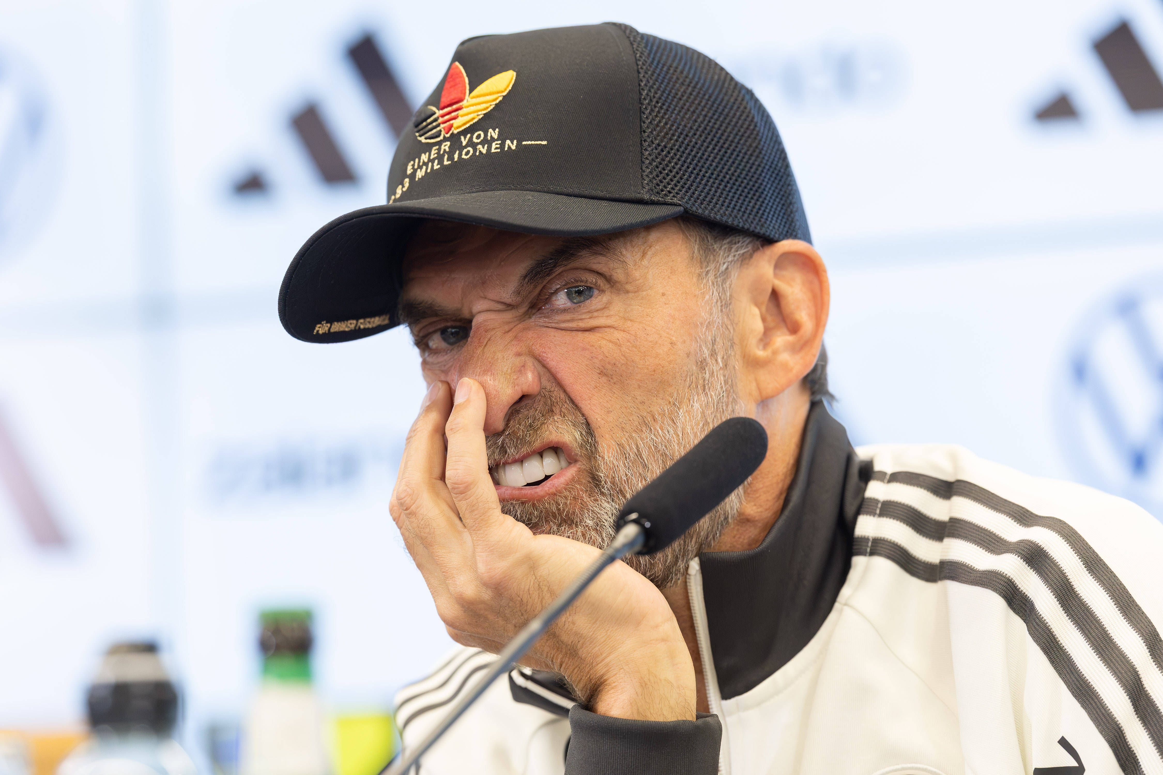Ein Mann mit Bart und Basecap kratzt sich mit Grimasse an der Nase bei einer Pressekonferenz.