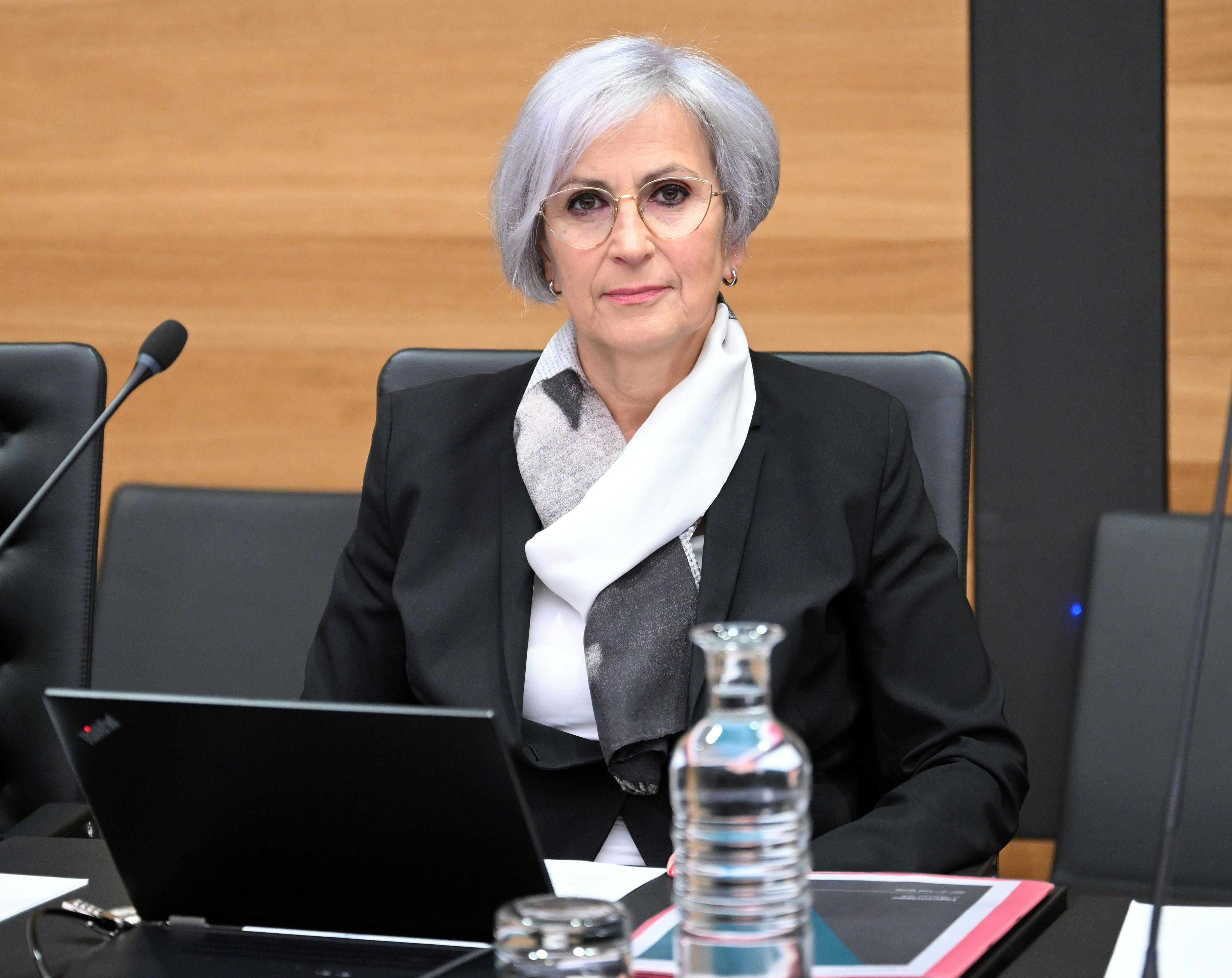 Eine Frau mit grauen Haaren sitzt im Anzug vor einem Laptop in einem Sitzungssaal.
