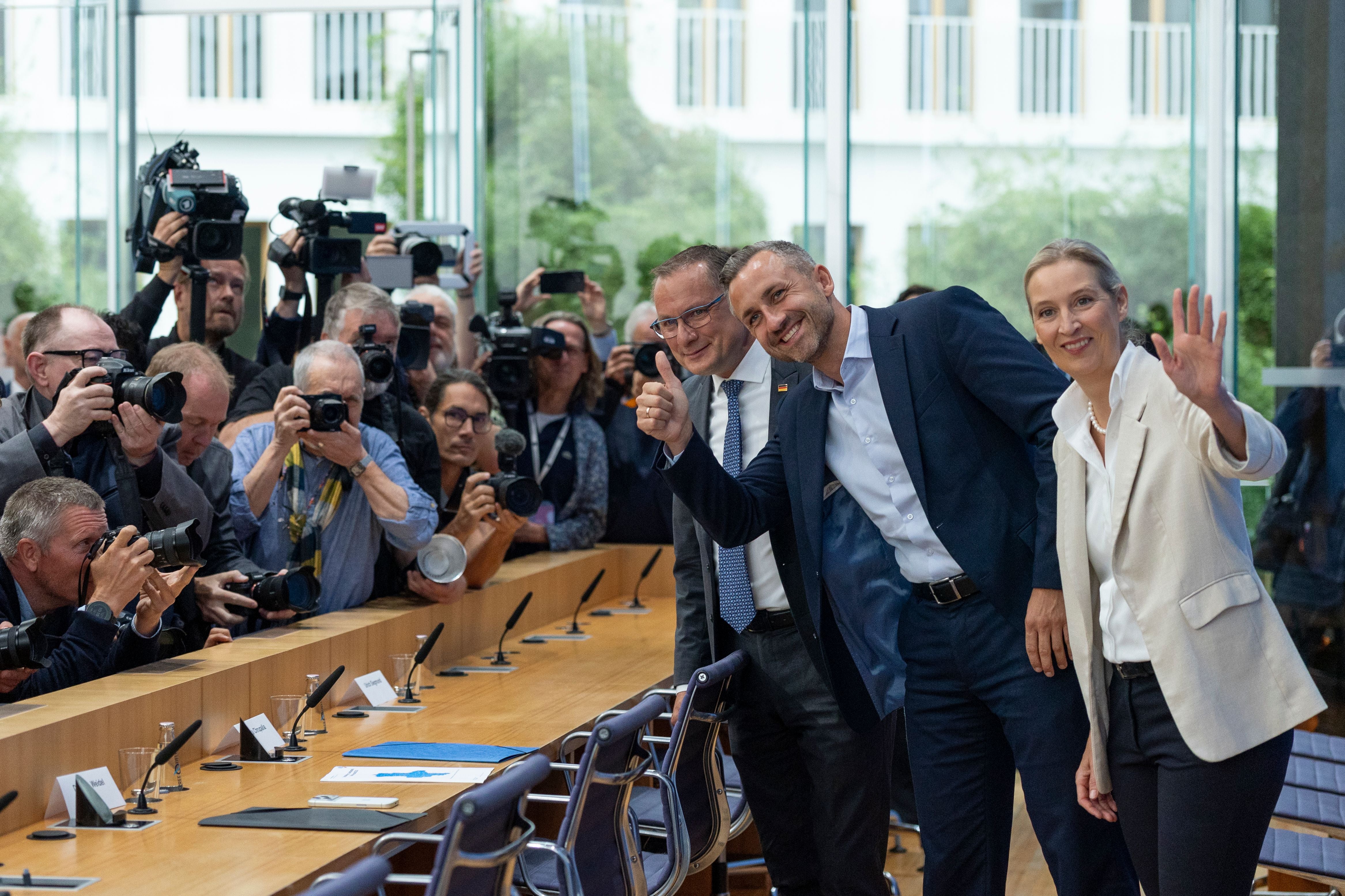 Drei Menschen vor einer Gruppe Fotografen, zwei lächeln und winken, einer zeigt den Daumen hoch.