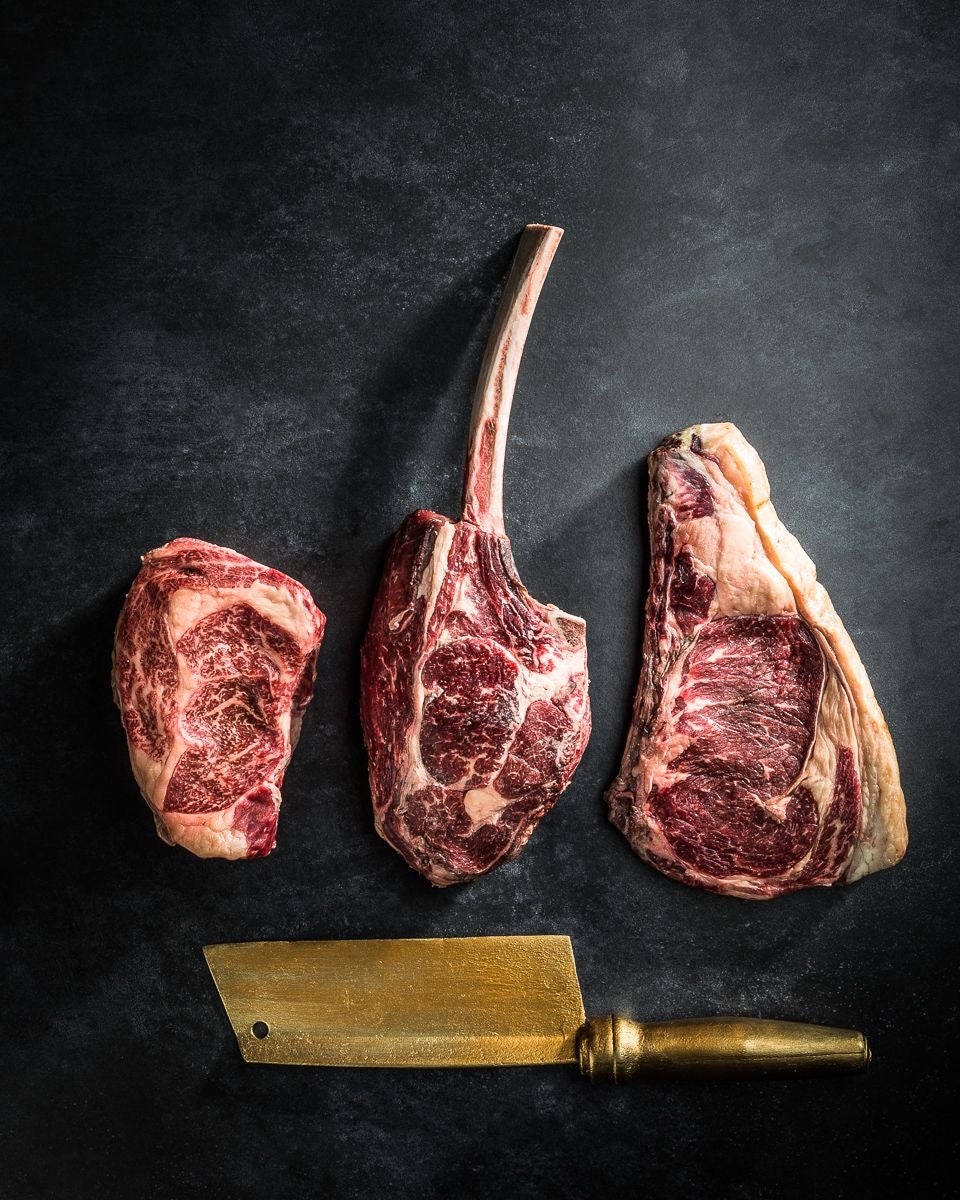 Drei verschiedene Rindersteaks und ein goldenes Hackmesser auf dunklem Untergrund.
