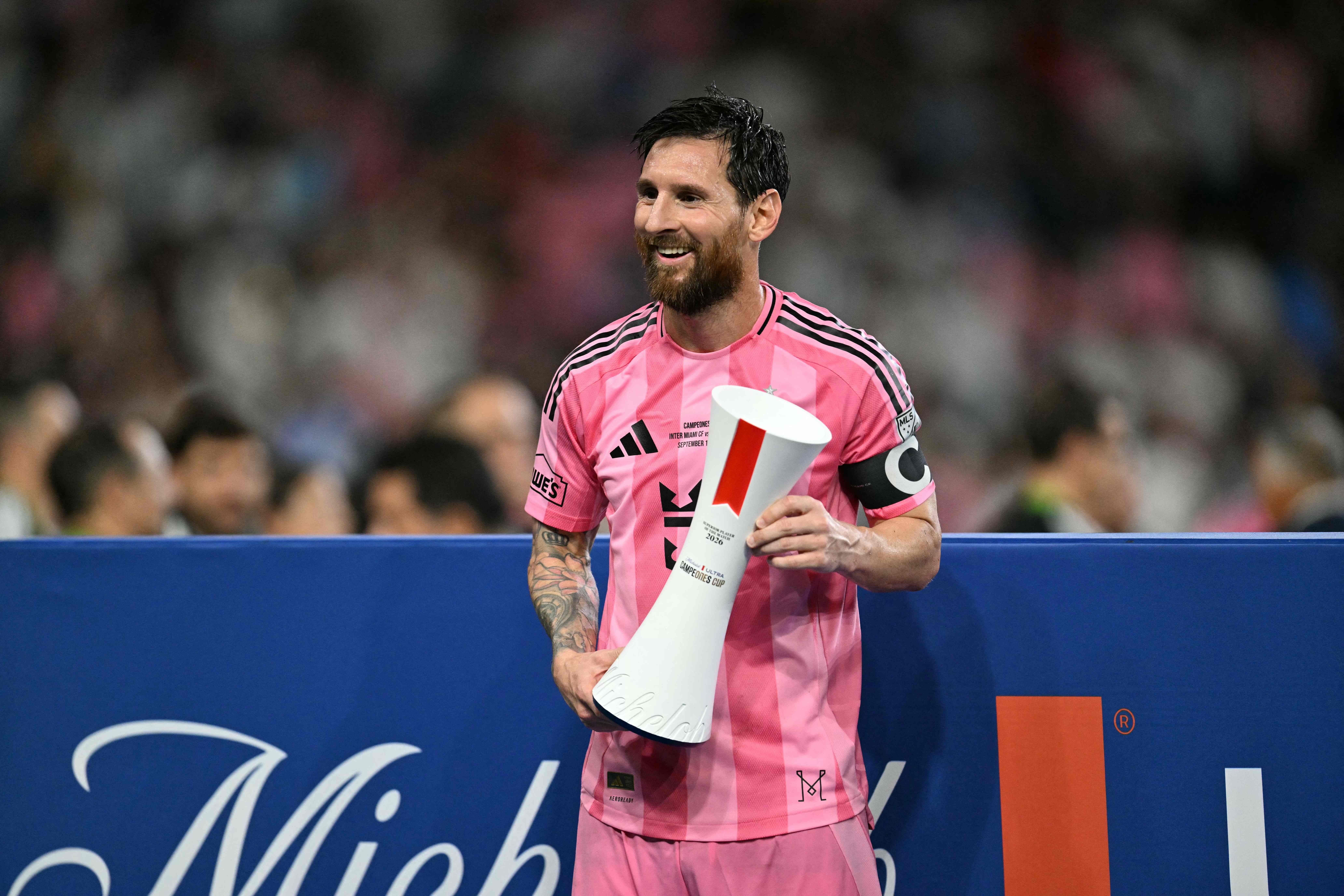 Ein Fußballspieler im pinken Trikot hält eine Trophäe und lächelt im Stadion.