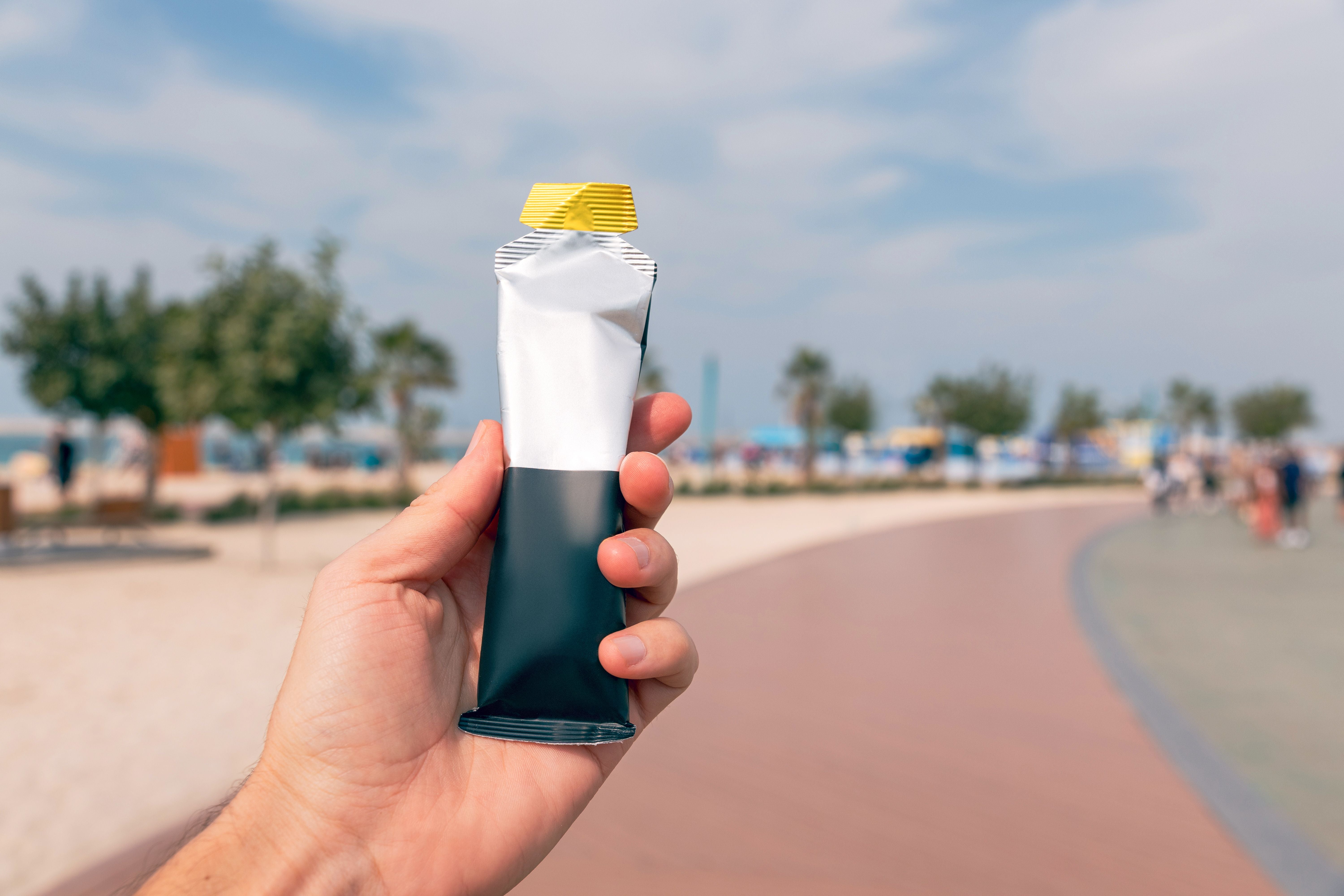 Hand hält Energiegel mit gelbem Deckel auf einer Promenade im Freien, unscharfer Hintergrund.
