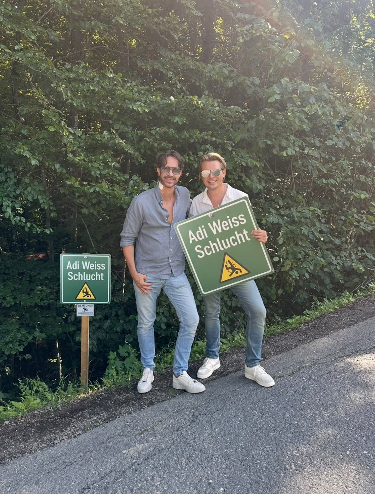 Zwei Männer posieren lächelnd mit einem Schild „Adi Weiss Schlucht“ am Waldrand.