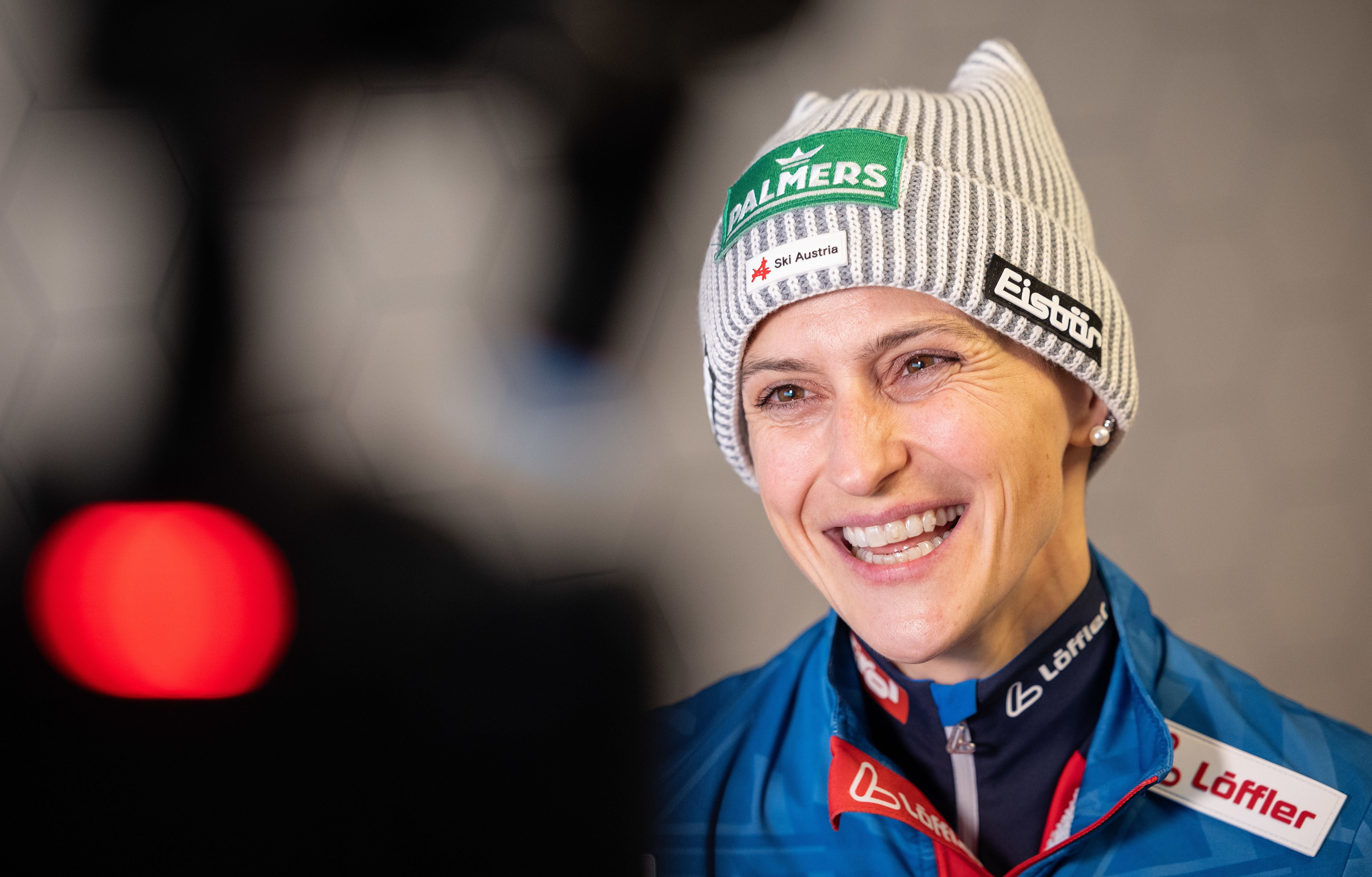 Skispringerin Eva Pinkelnig lächelt im ÖSV-Outfit mit grauer Eisbähr-Mütze vor einer Kamera.