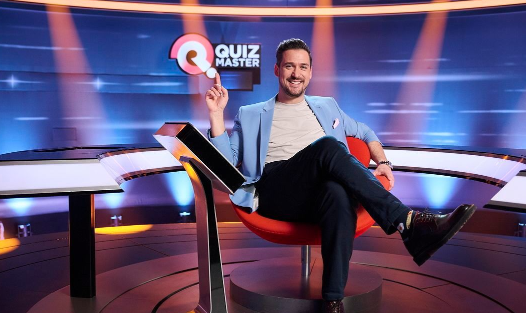 Ein Mann sitzt lächelnd in einem Fernsehstudio auf einem roten Stuhl, hinter ihm steht "Quizmaster".