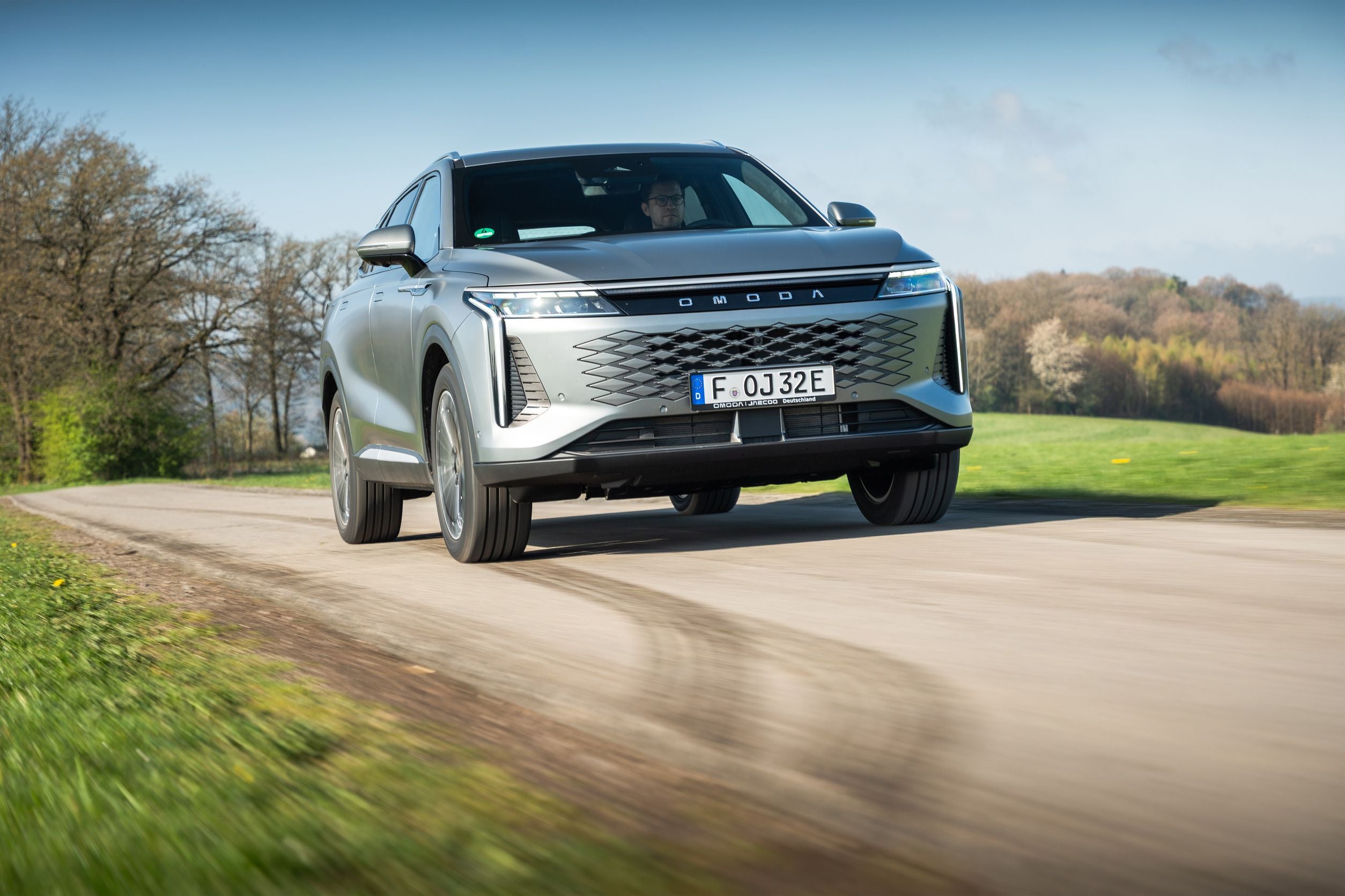 Silberner Omoda SUV fährt auf einer Landstraße durch grüne Landschaft mit Bäumen.