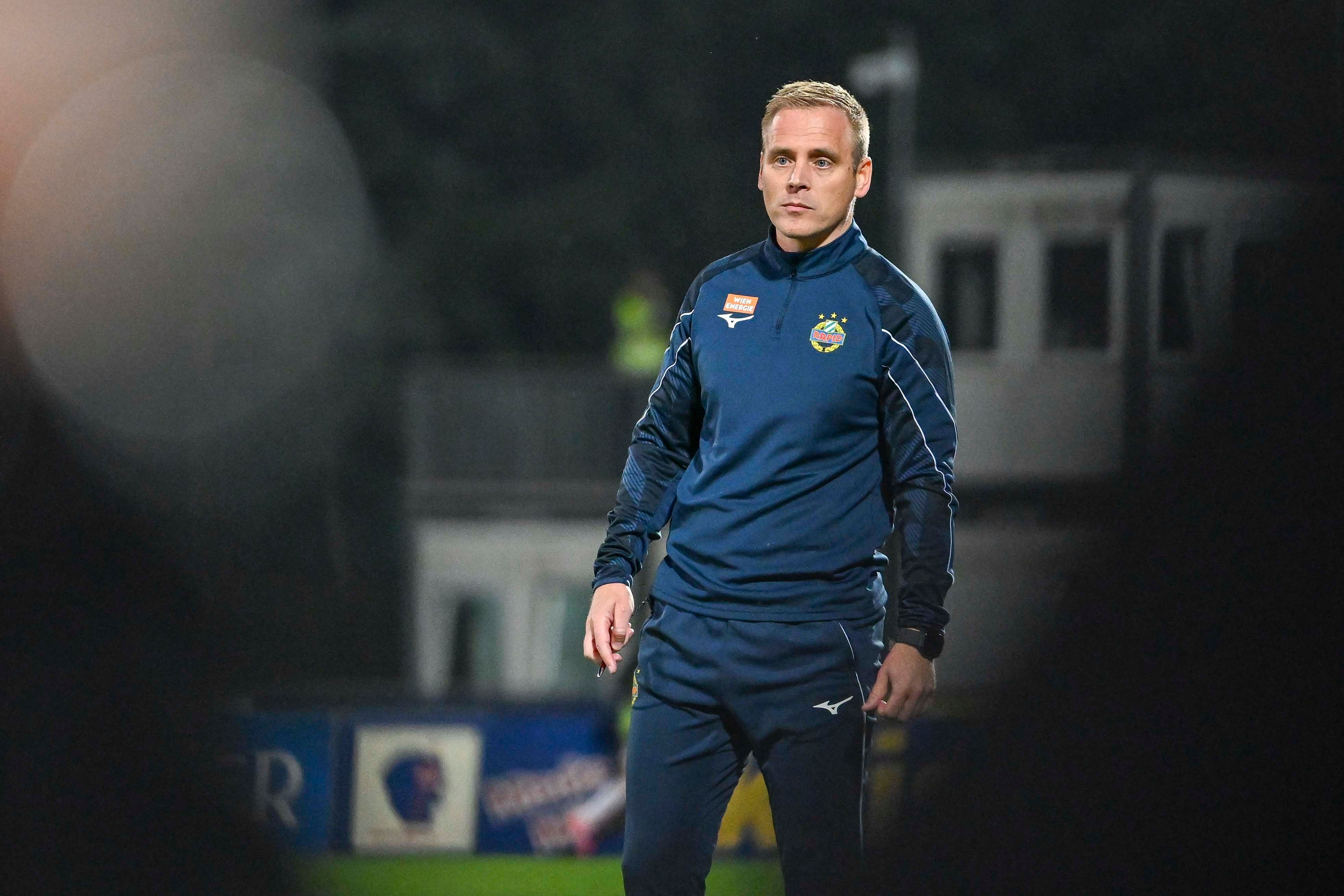 Ein Fußballtrainer im Trainingsanzug steht auf einem Spielfeld am Abend.