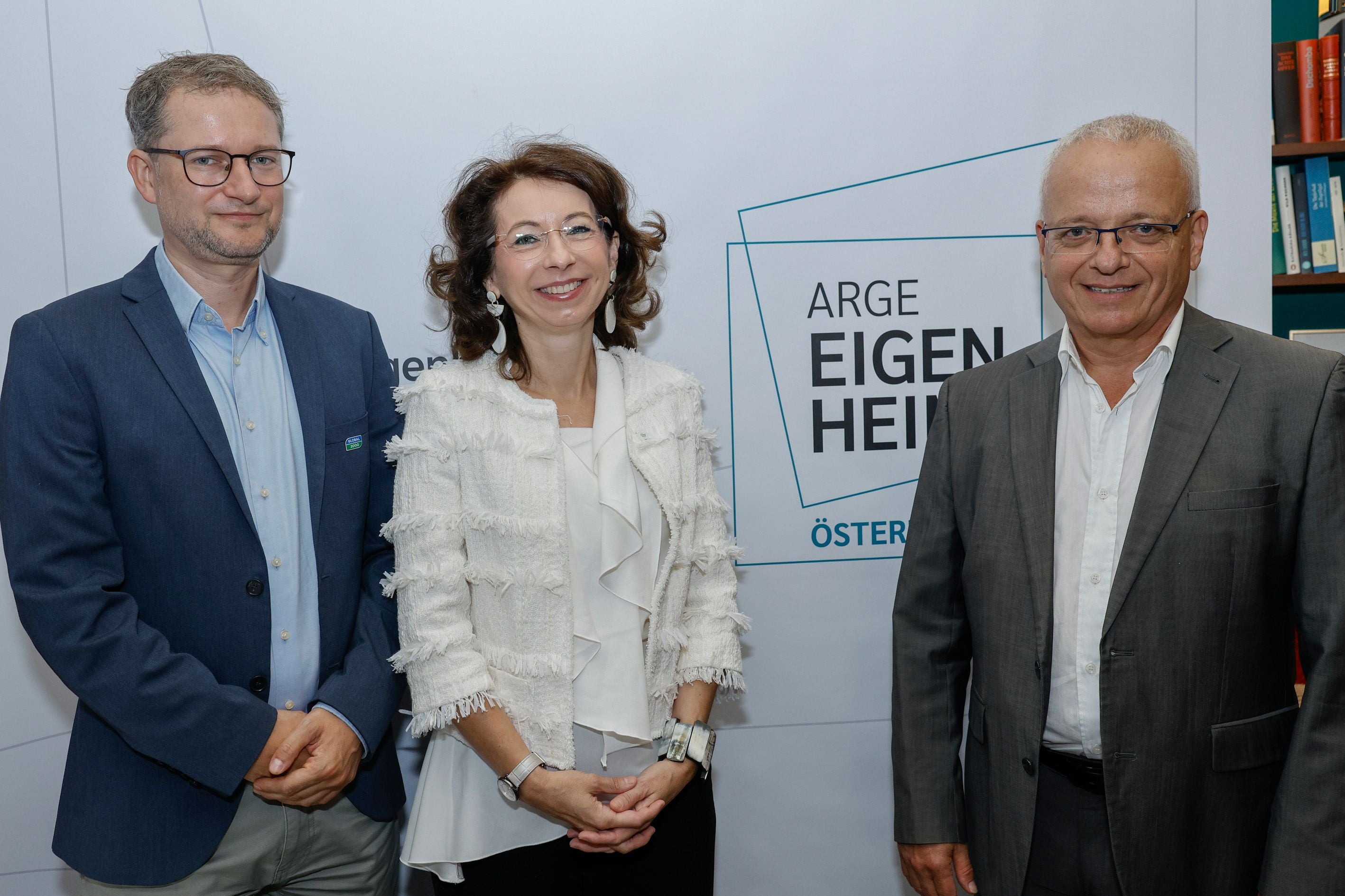 Drei Personen in Businesskleidung stehen vor einem ARGE Eigenheim Hintergrund und lächeln.