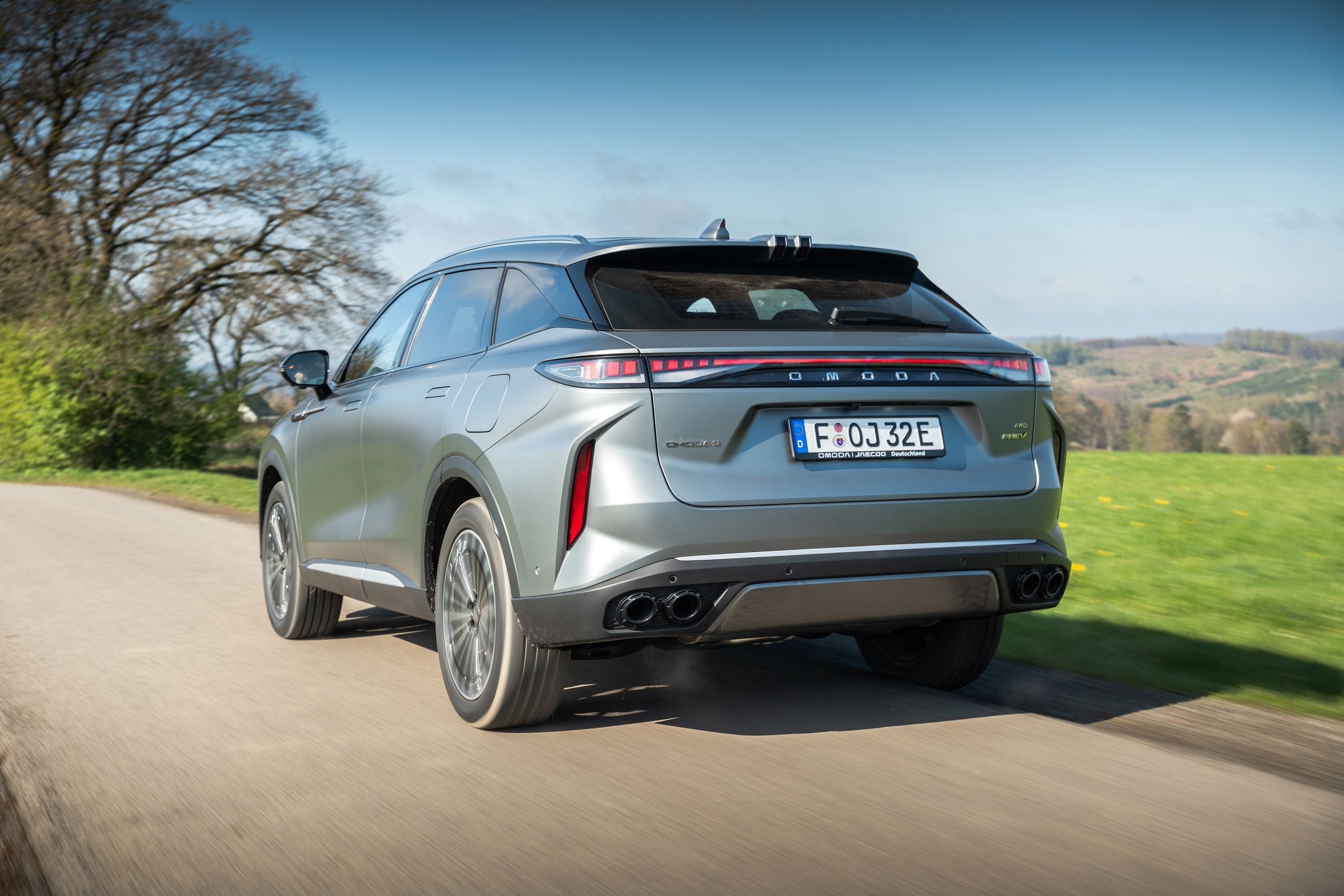 Silberner SUV fährt auf einer Landstraße, im Hintergrund grüne Wiesen und Bäume.