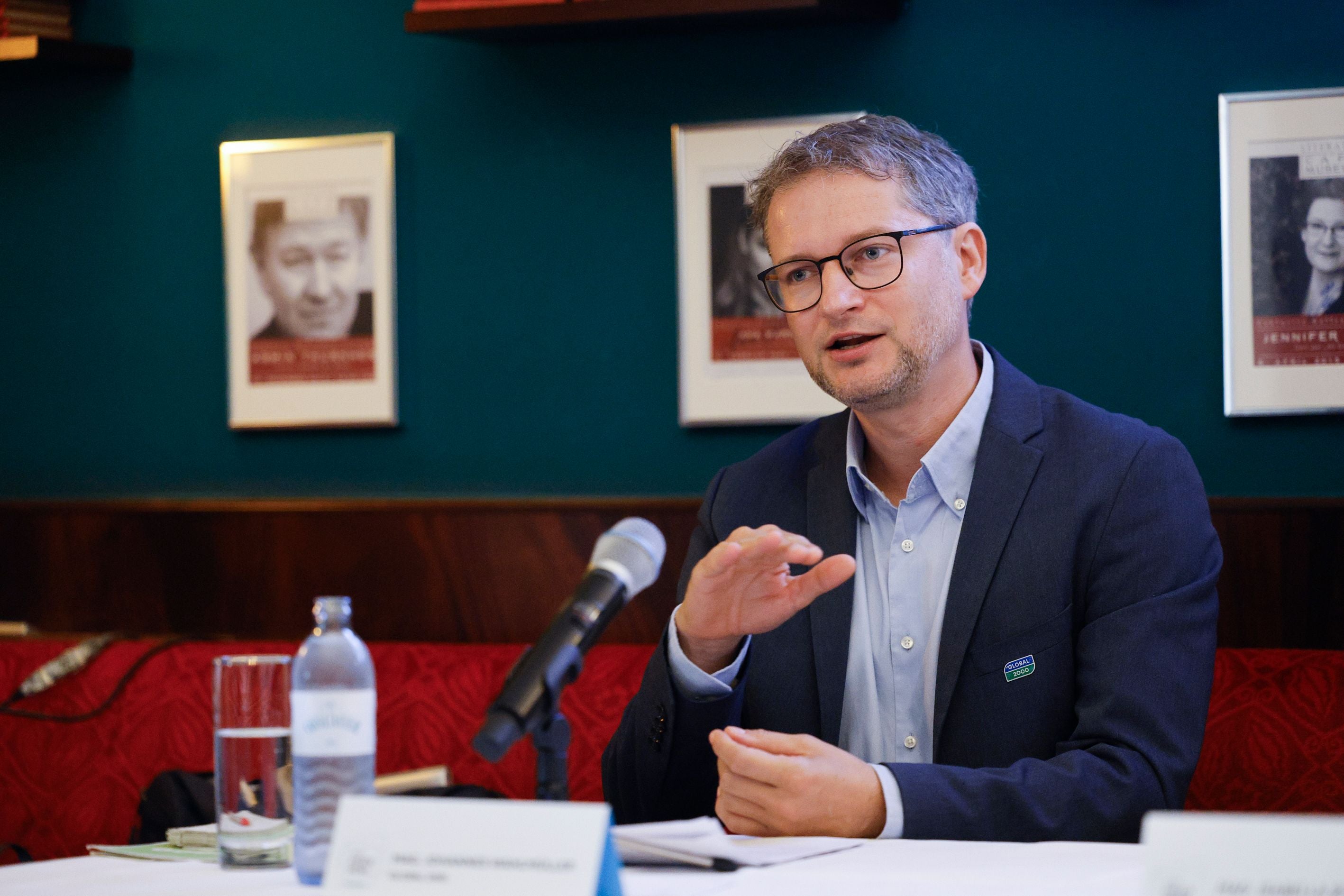 Ein Mann im Anzug spricht bei einer Pressekonferenz, ein Mikrofon und Wasserflasche vor ihm.