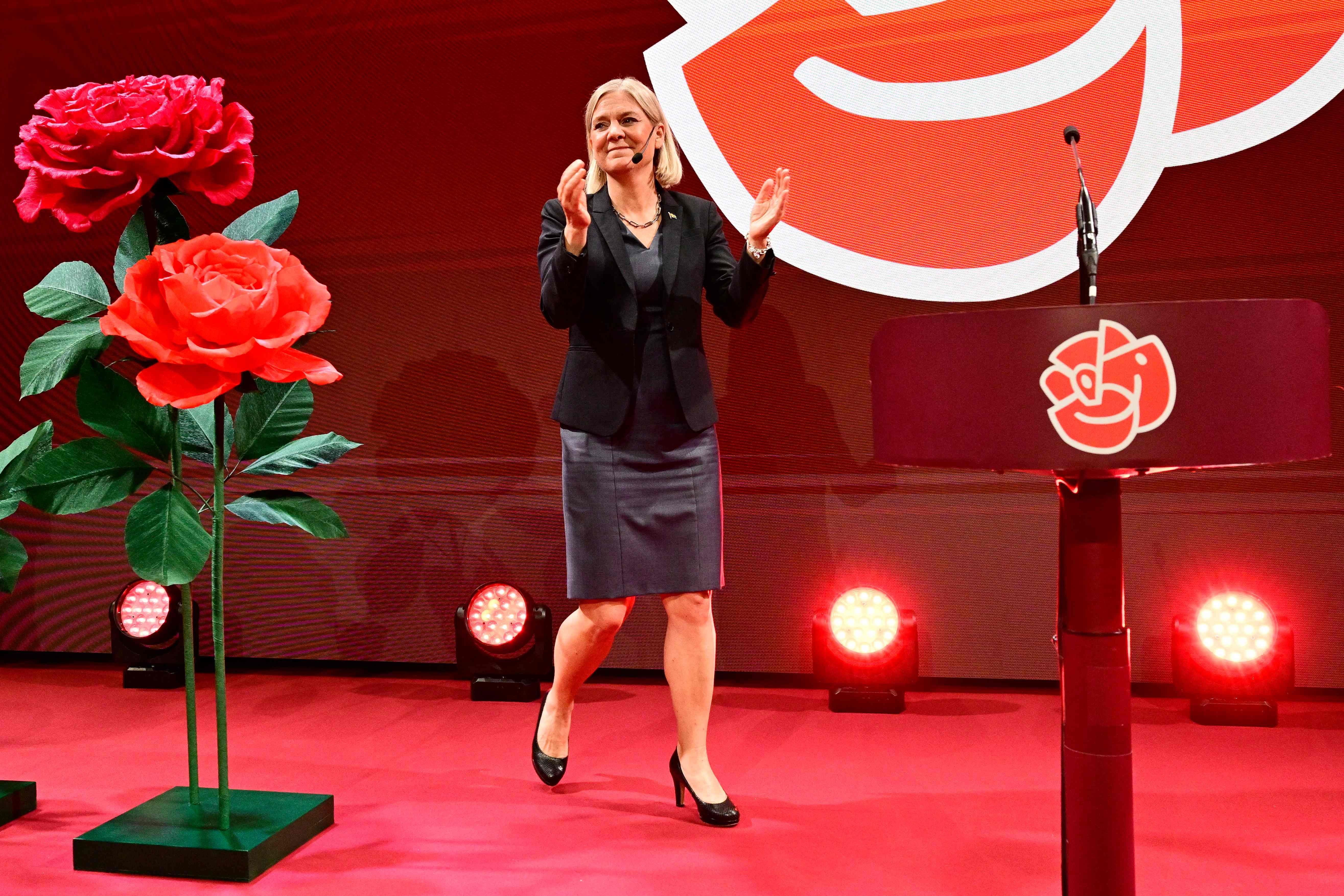 Eine Frau steht auf einer rot beleuchteten Bühne neben großen roten Rosen und einem Rednerpult.