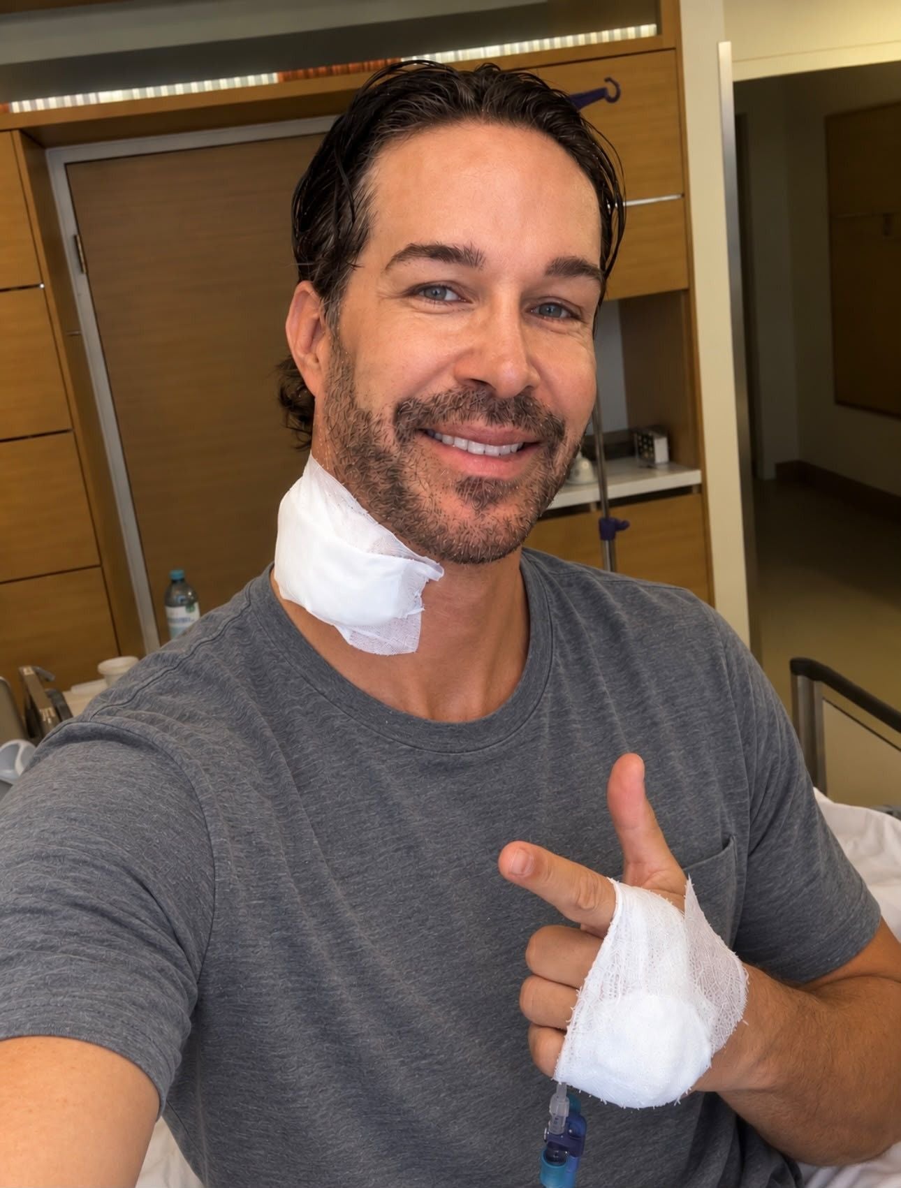 Ein lächelnder Mann mit Verbänden am Hals und an der Hand sitzt in einem Krankenhauszimmer.