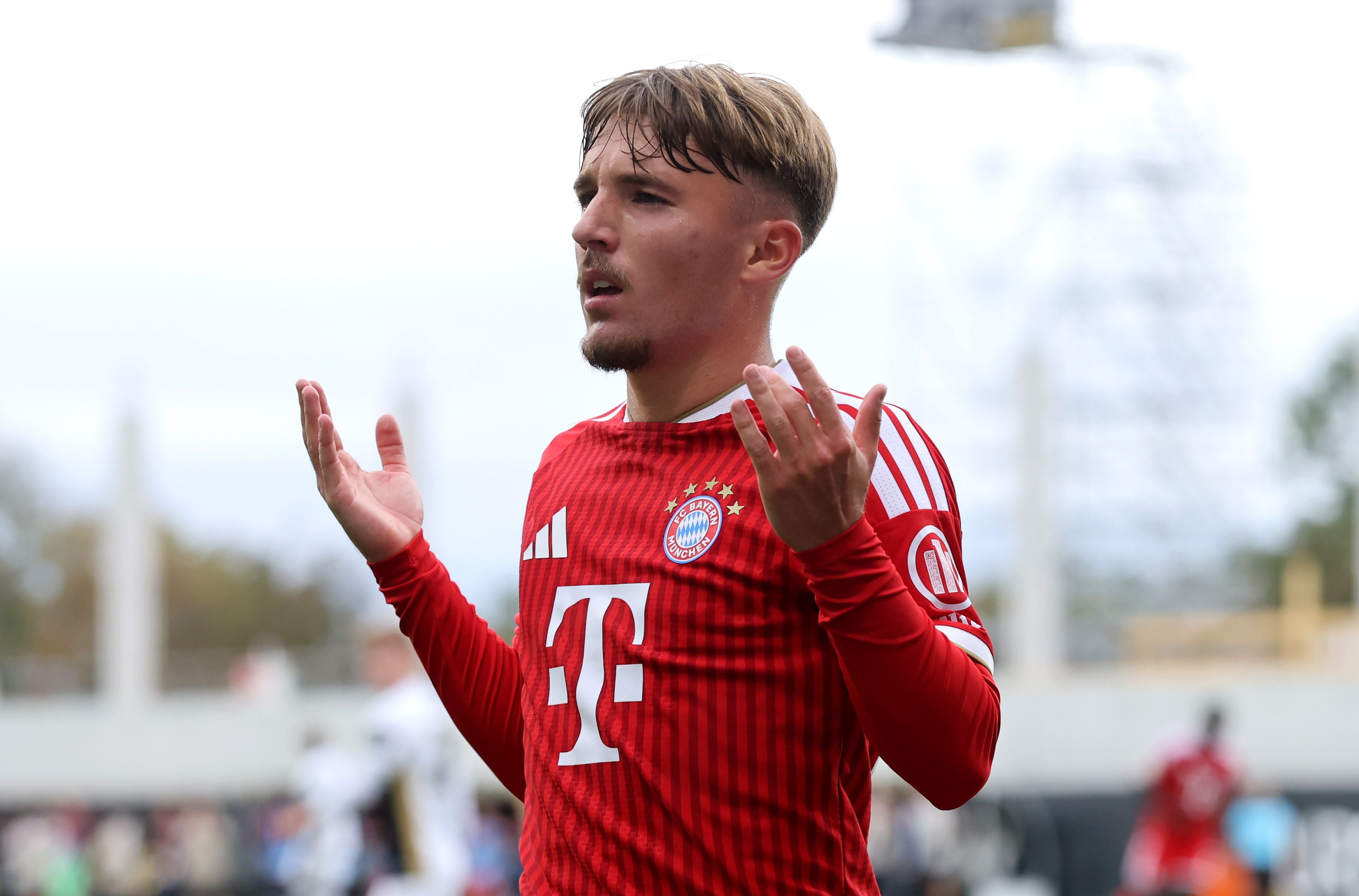 Ein Fußballspieler im roten Bayern-Trikot hebt fragend die Arme während eines Spiels.