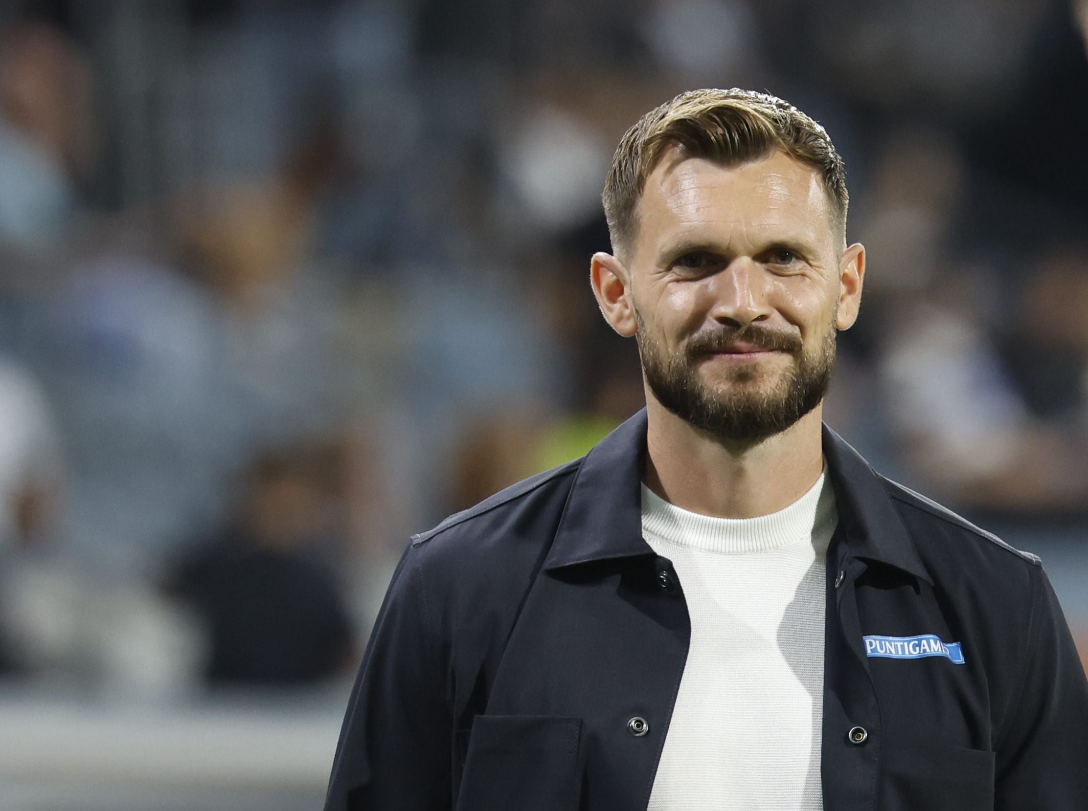 Ein Mann mit Bart und kurzem Haar lächelt im Stadion, trägt dunkle Jacke und weißes Shirt.