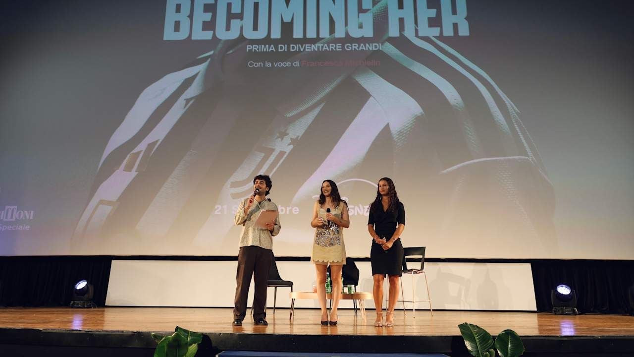 Drei Personen stehen auf einer Bühne vor einer Leinwand mit dem Schriftzug "Becoming Her".