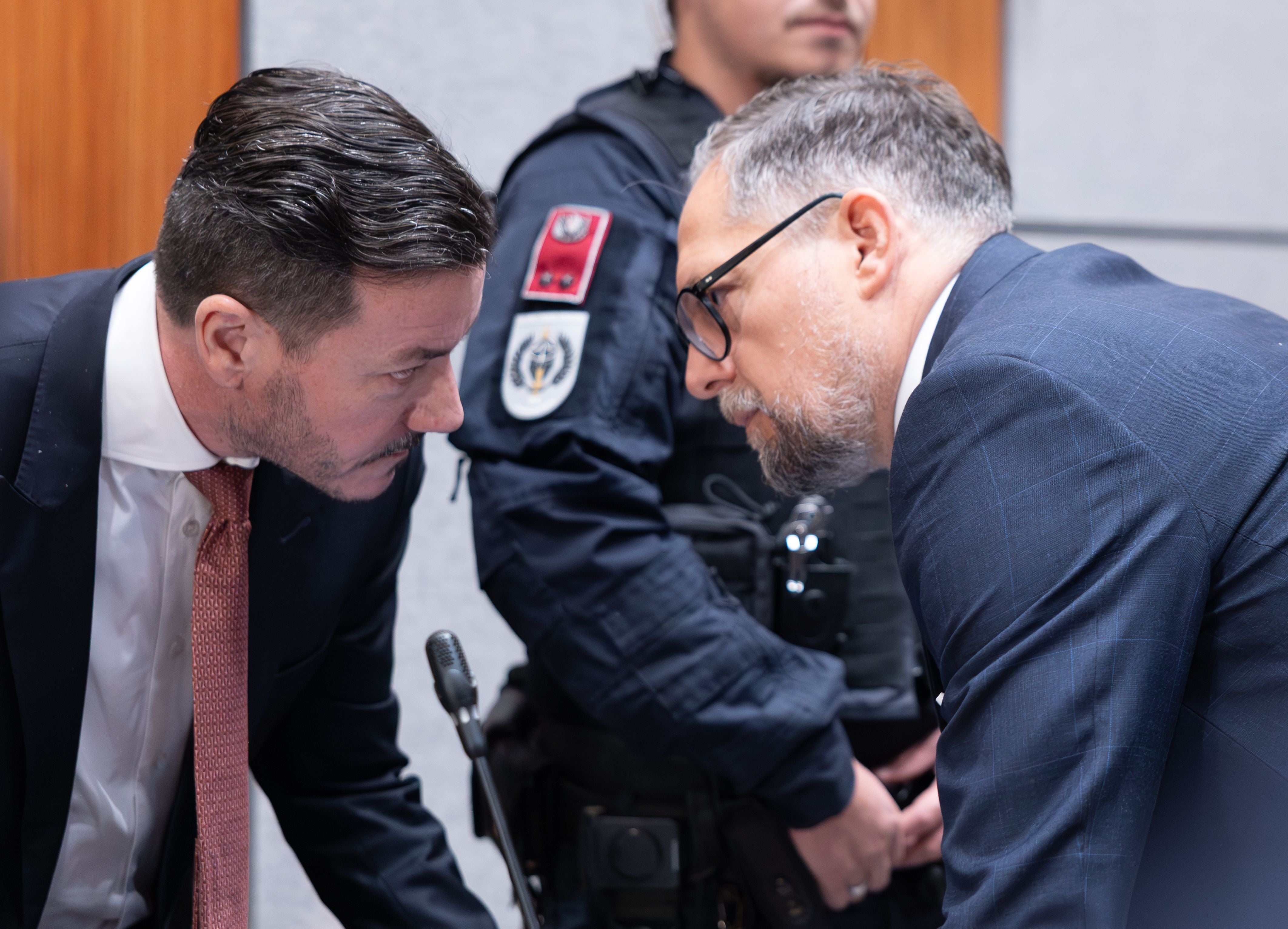 Zwei Männer im Anzug sprechen sich vor Gericht vertraulich, ein Polizist steht dahinter.