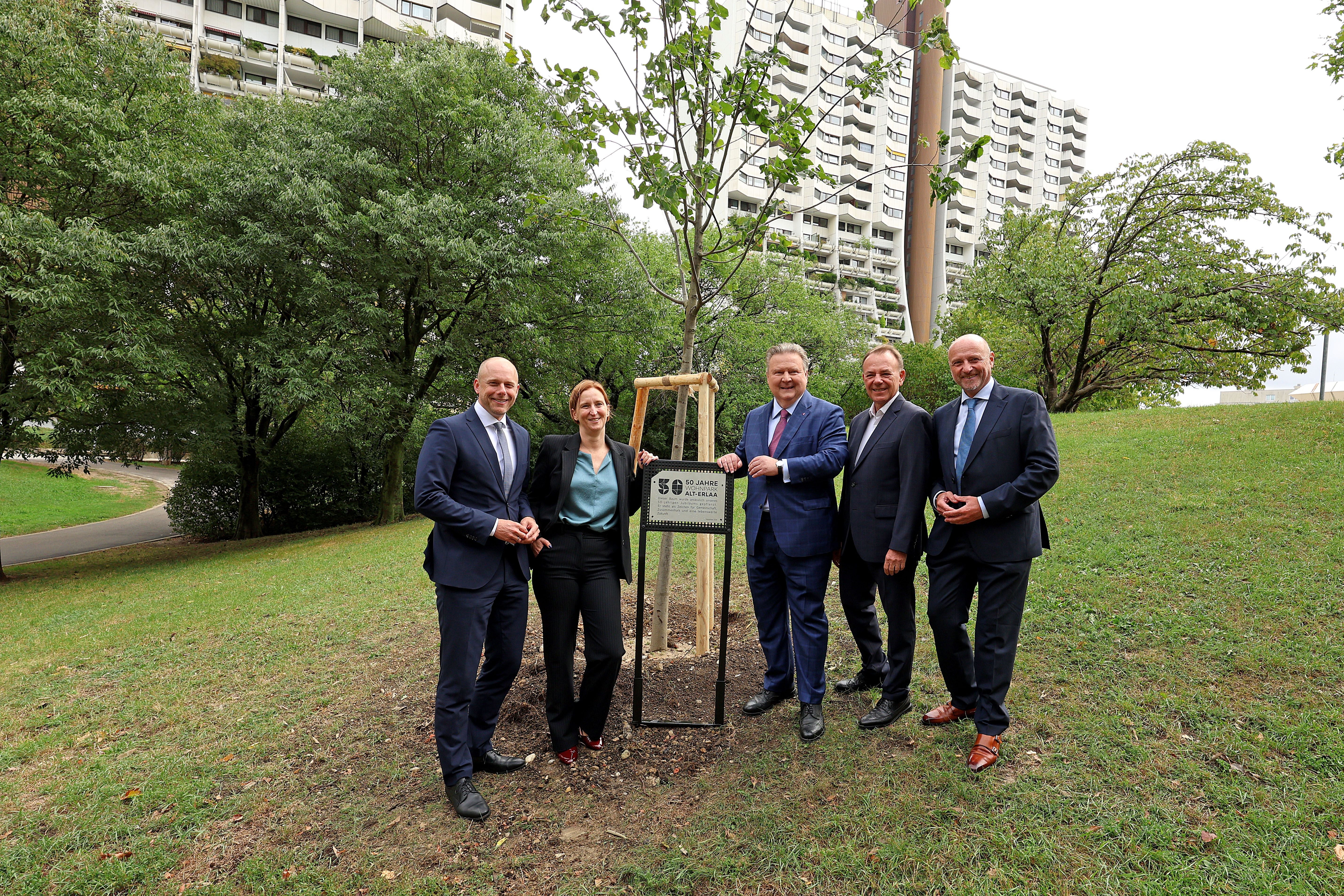 Fünf Personen stehen bei einem neu gepflanzten Baum mit Schild auf einer Wiese vor Wohnhäusern.