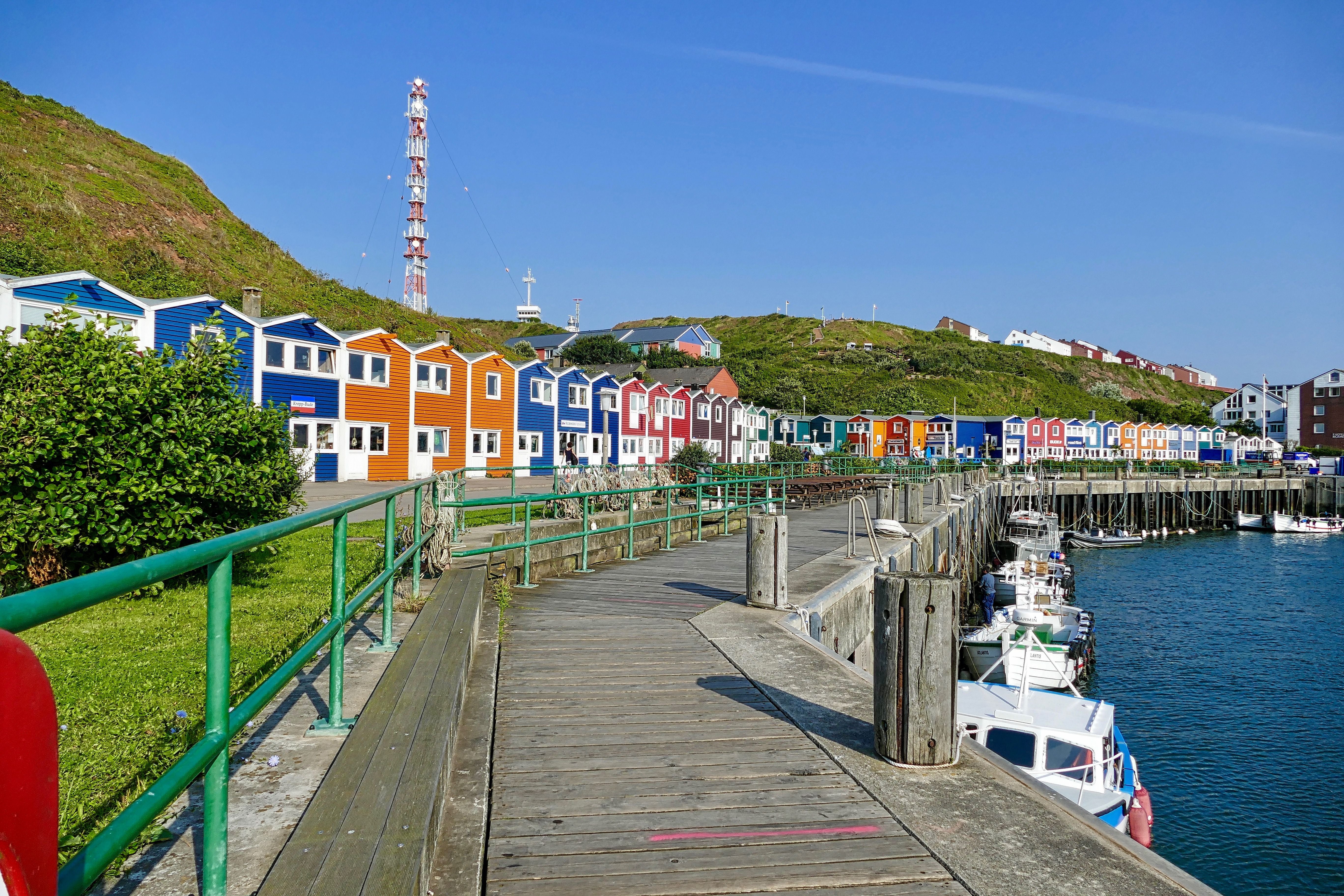Bunte Häuser am Hafen von Helgoland, davor Boote im Wasser und ein Holzsteg.