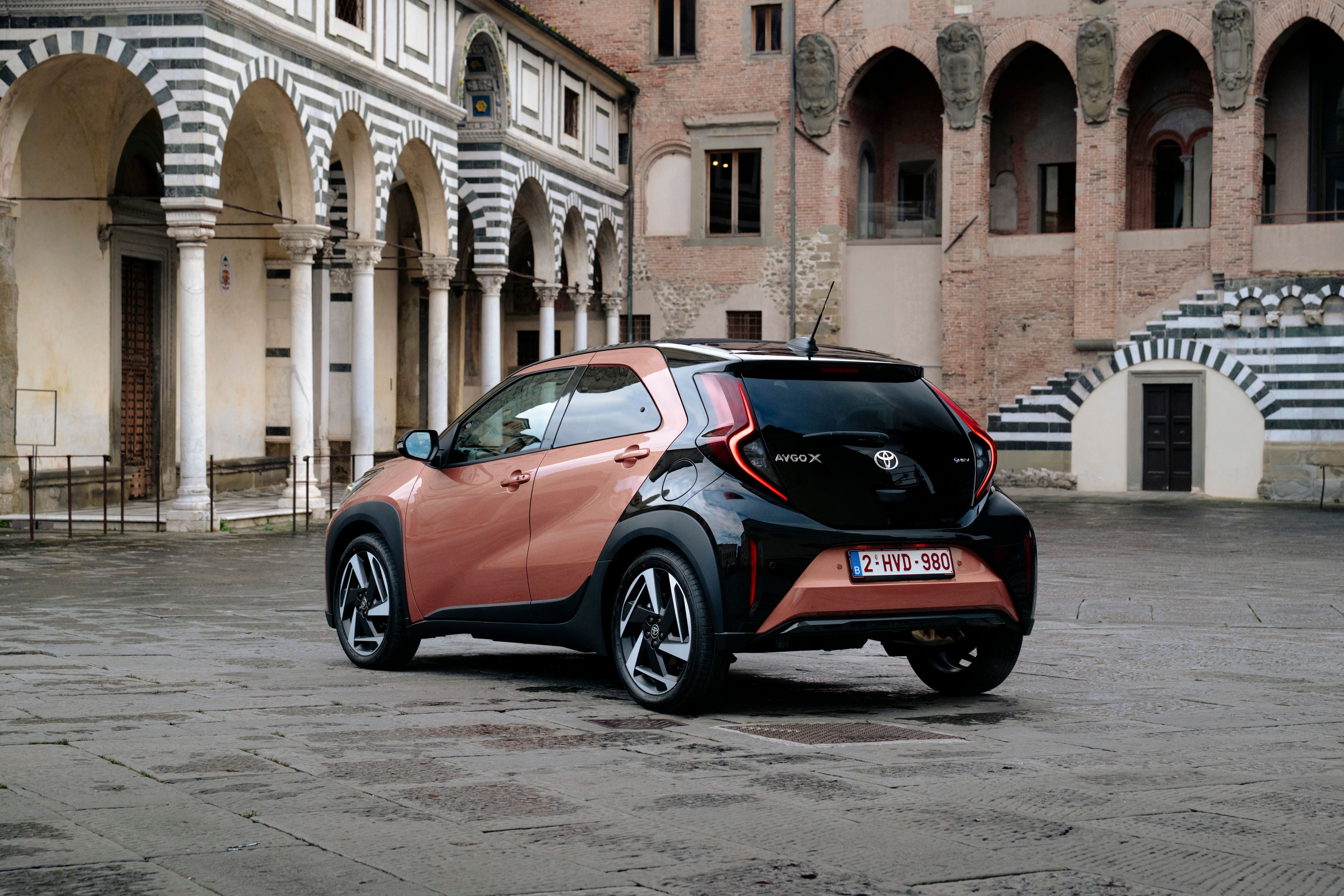 Ein orange-schwarzer Toyota Aygo X steht vor historischen Gebäuden mit Arkaden und Ziegelmauern.