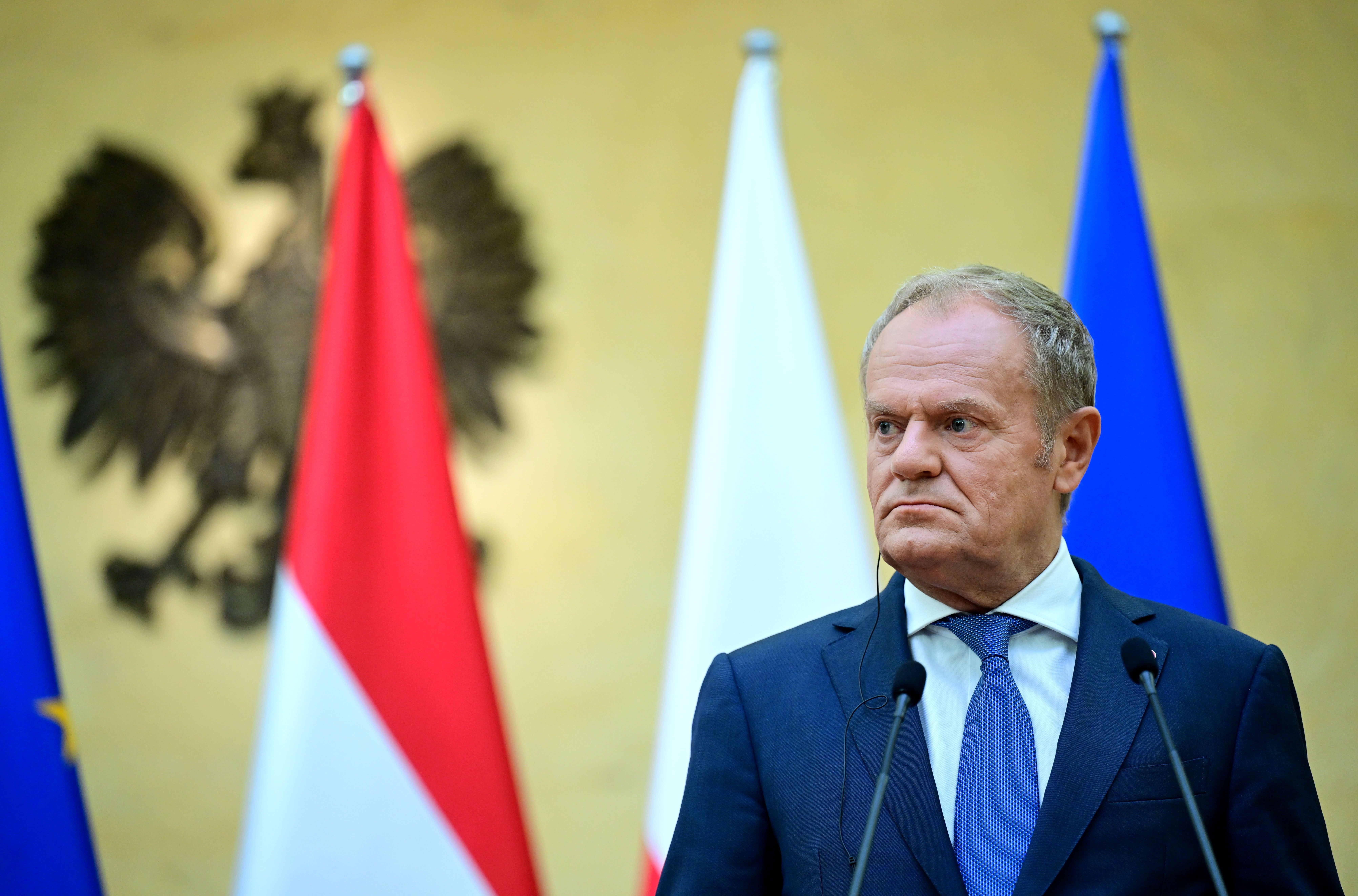Ein Mann im Anzug steht vor Mikrofonen und Flaggen bei einer Pressekonferenz.
