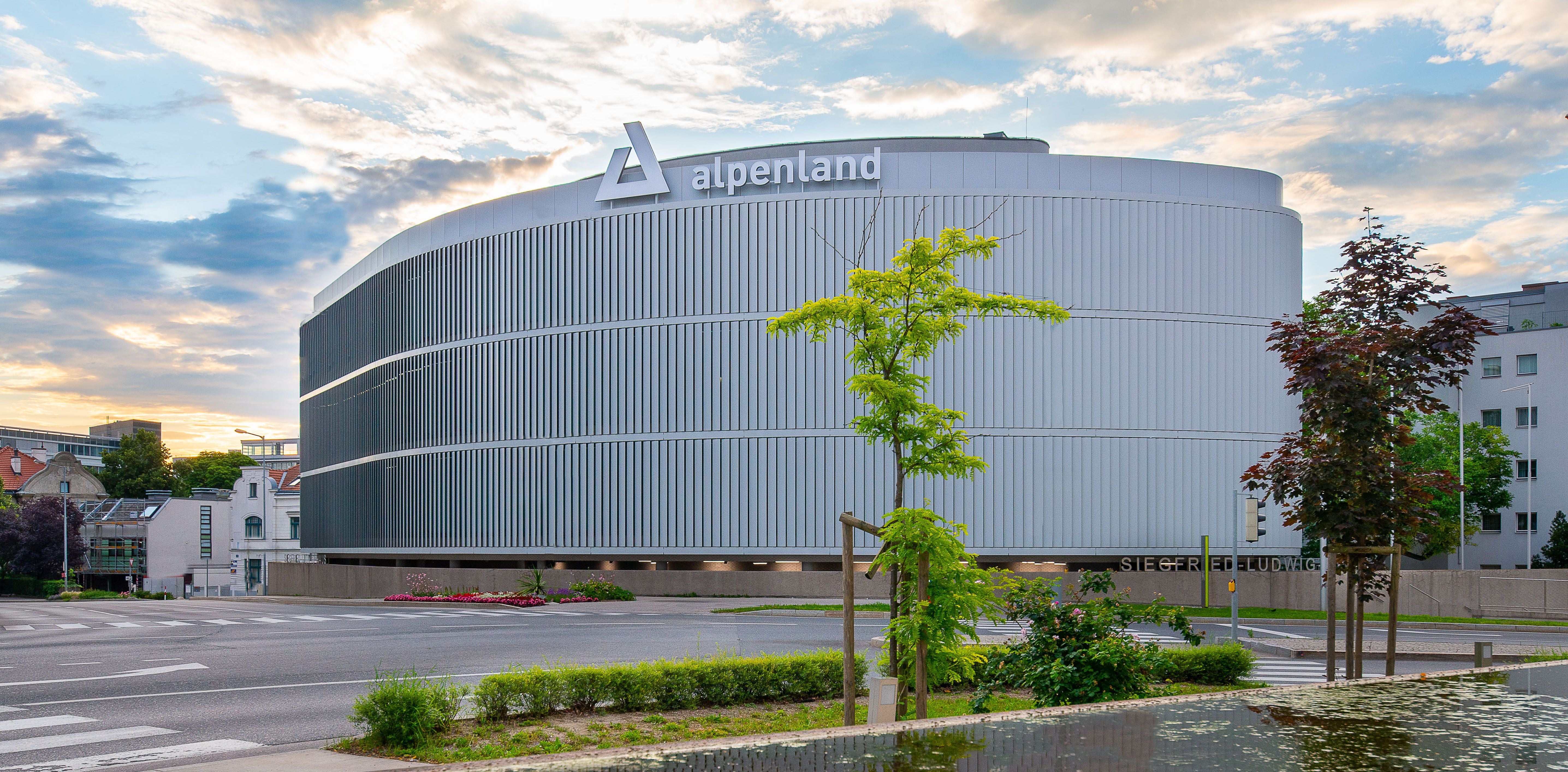 Modernes Bürogebäude der Alpenland Zentrale mit runder Fassade und Bäumen im Vordergrund.