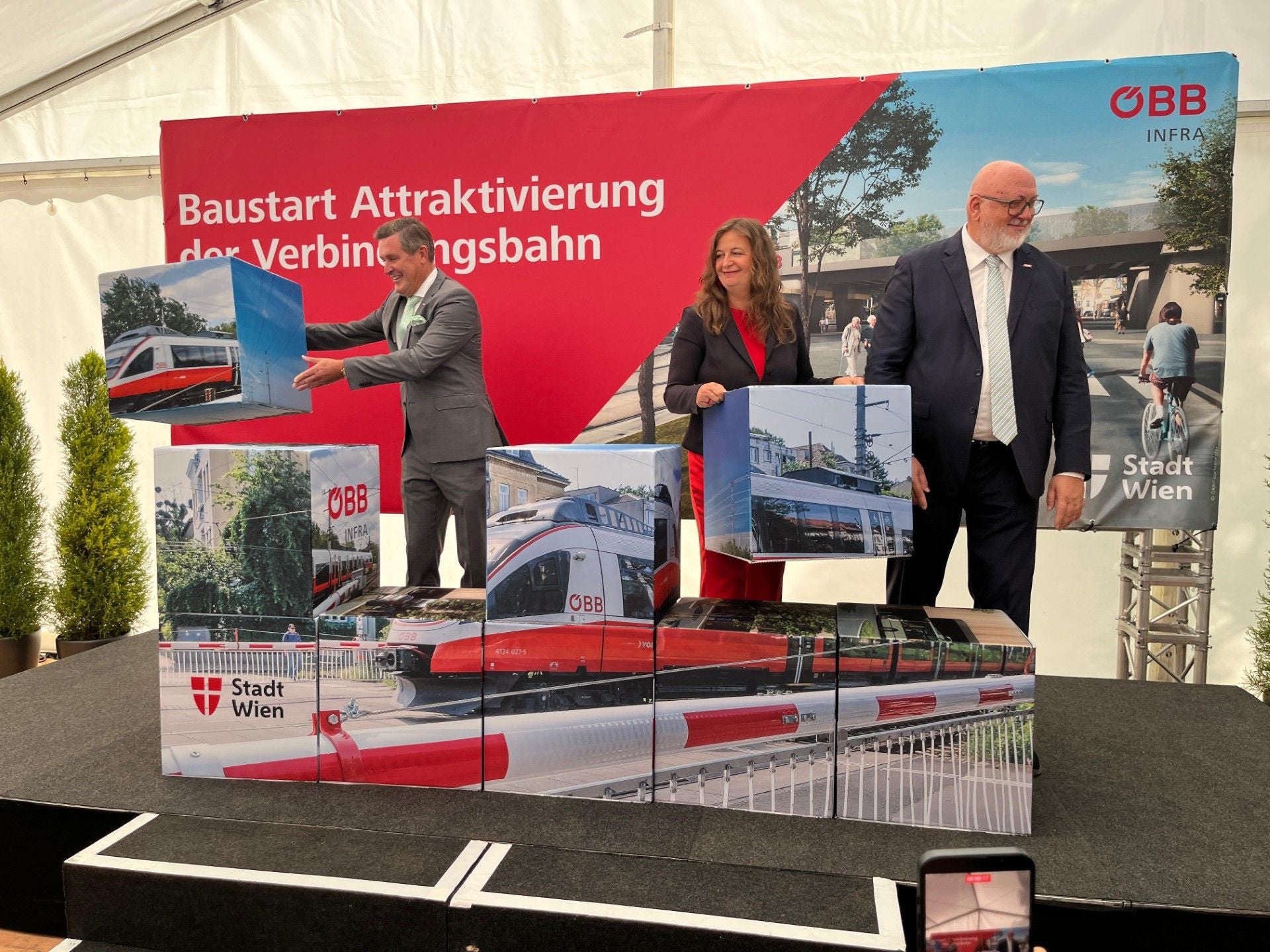 Drei Personen bauen auf einer Bühne aus Würfeln ein Bild eines ÖBB-Zugs vor Werbeplakaten zusammen.