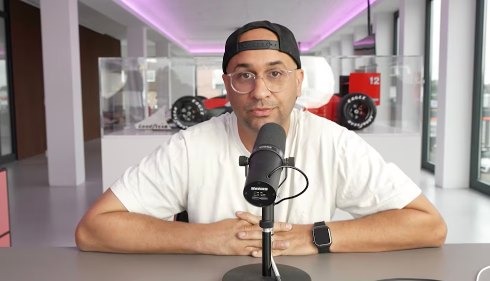Mann mit Brille und Cap spricht in Mikrofon, im Hintergrund ein Formel-1-Auto-Modell.
