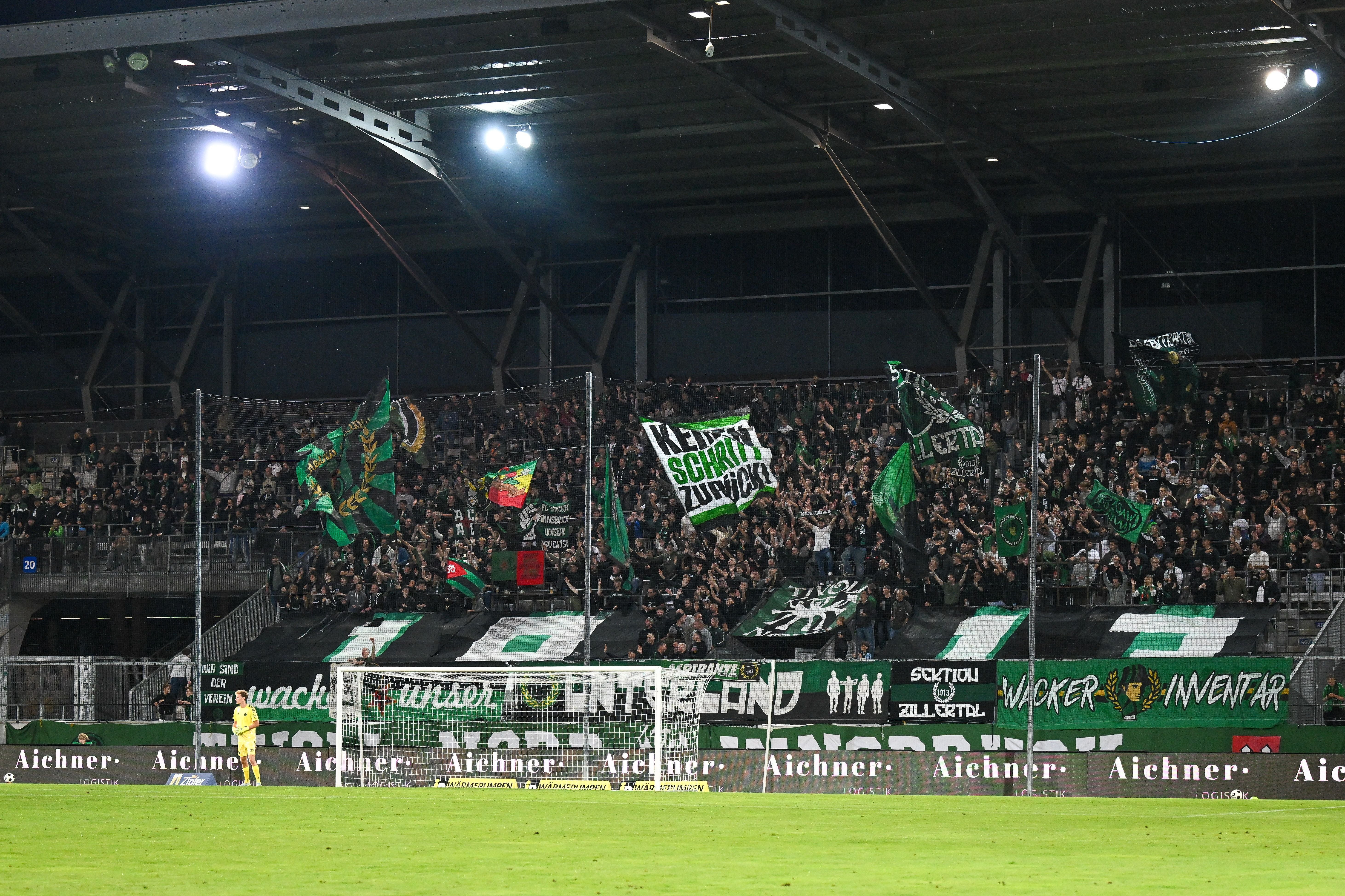 Fans von Wacker Innsbruck schwenken grüne und schwarze Fahnen im Stadion hinter dem Tor.