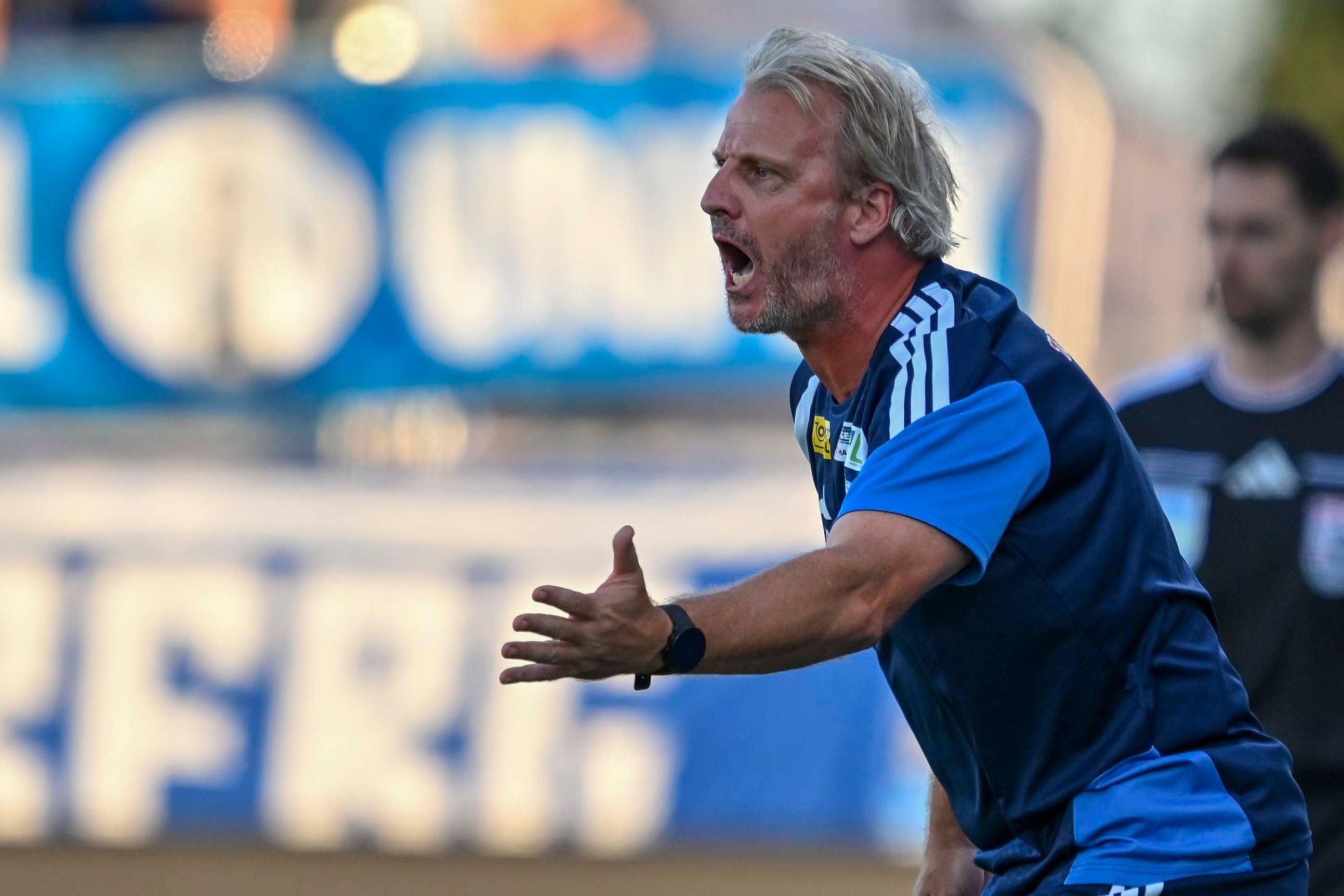 Ein Trainer in blauer Sportkleidung gestikuliert lautstark am Spielfeldrand.