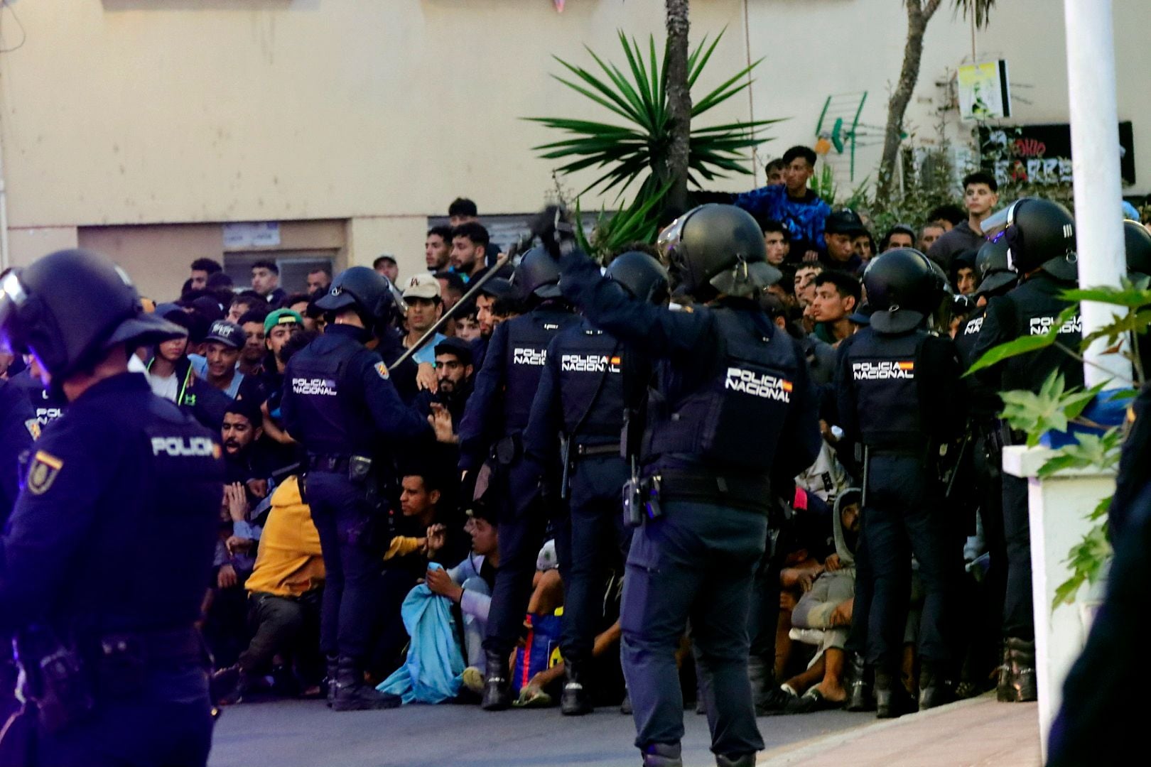 Polizisten in Schutzausrüstung überwachen eine große Gruppe sitzender Migranten im Freien.