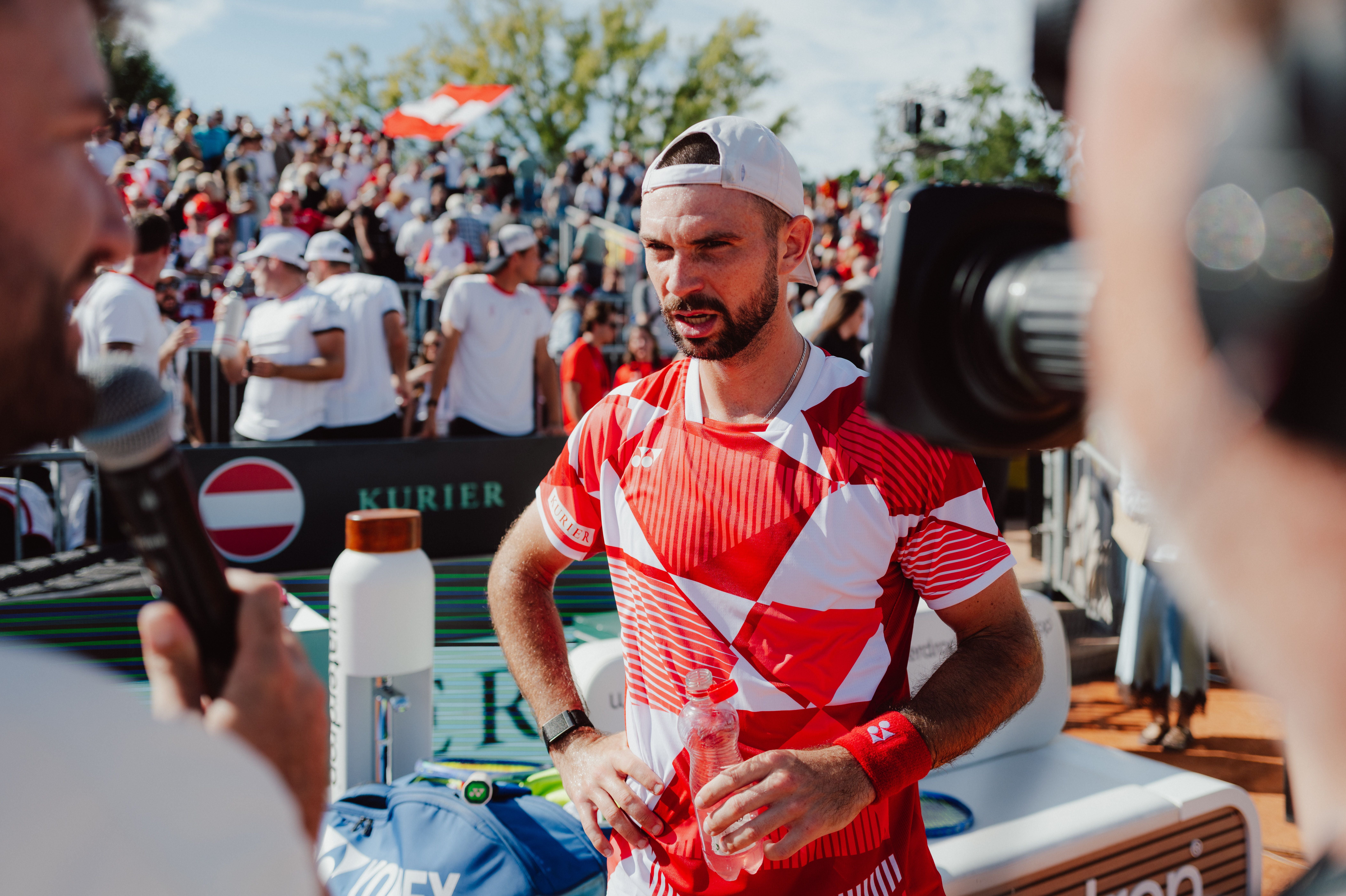 Jurij Rodionov gibt im rot-weiß gemusterten Trikot mit verkehrt getragener Kappe ein Interview auf dem Tennisplatz.