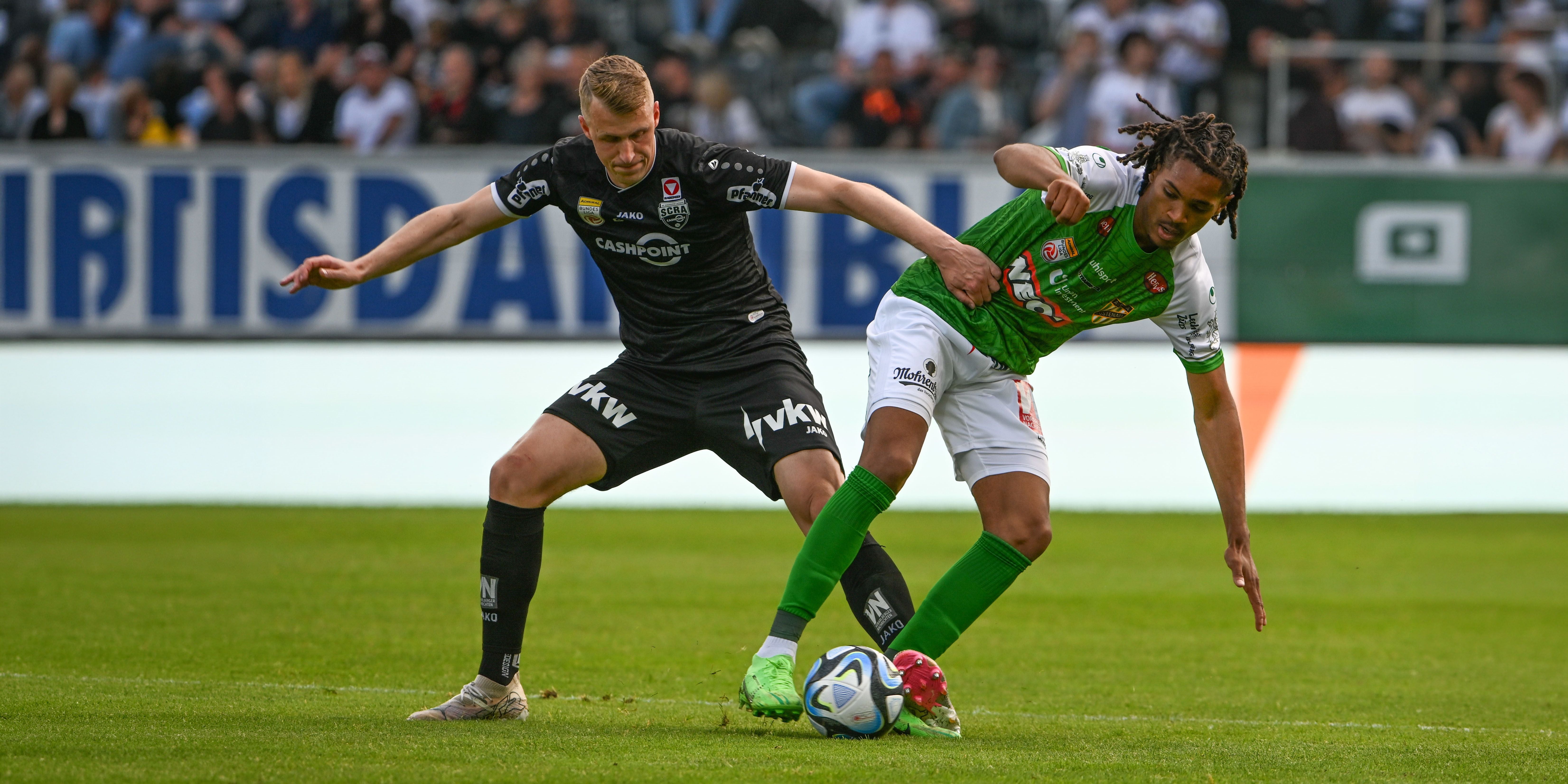 Lukas Jäger im schwarzen SCR-Altach-Trikot kämpft mit breiter Armhaltung im Zweikampf gegen einen Spieler von Austria Lustenau im grün-weißen Dress um den Ball.