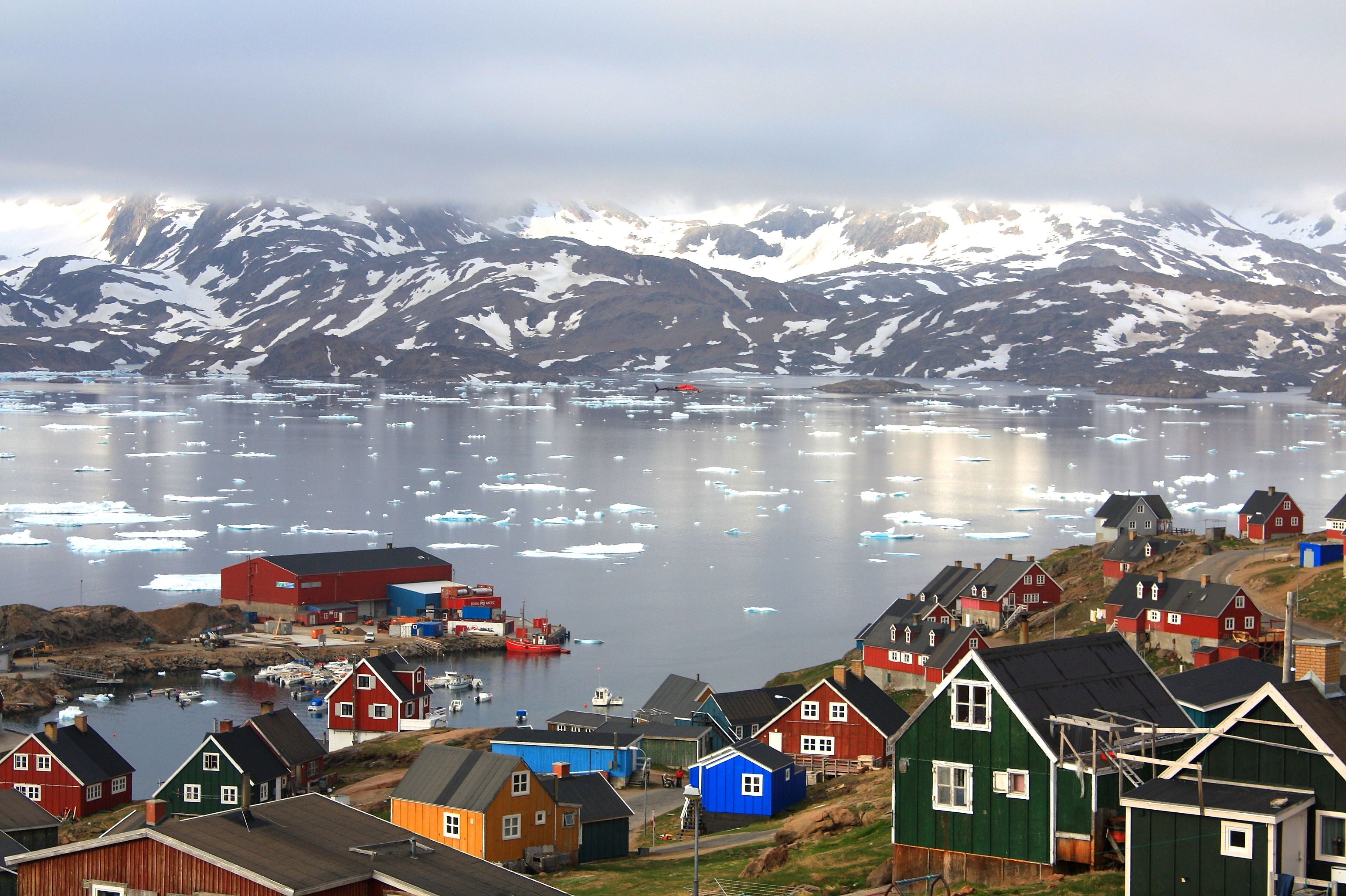 Bunte Häuser in Tasiilaq, Ostgrönland, mit Hafen, Eisbergen und verschneiten Bergen im Hintergrund.