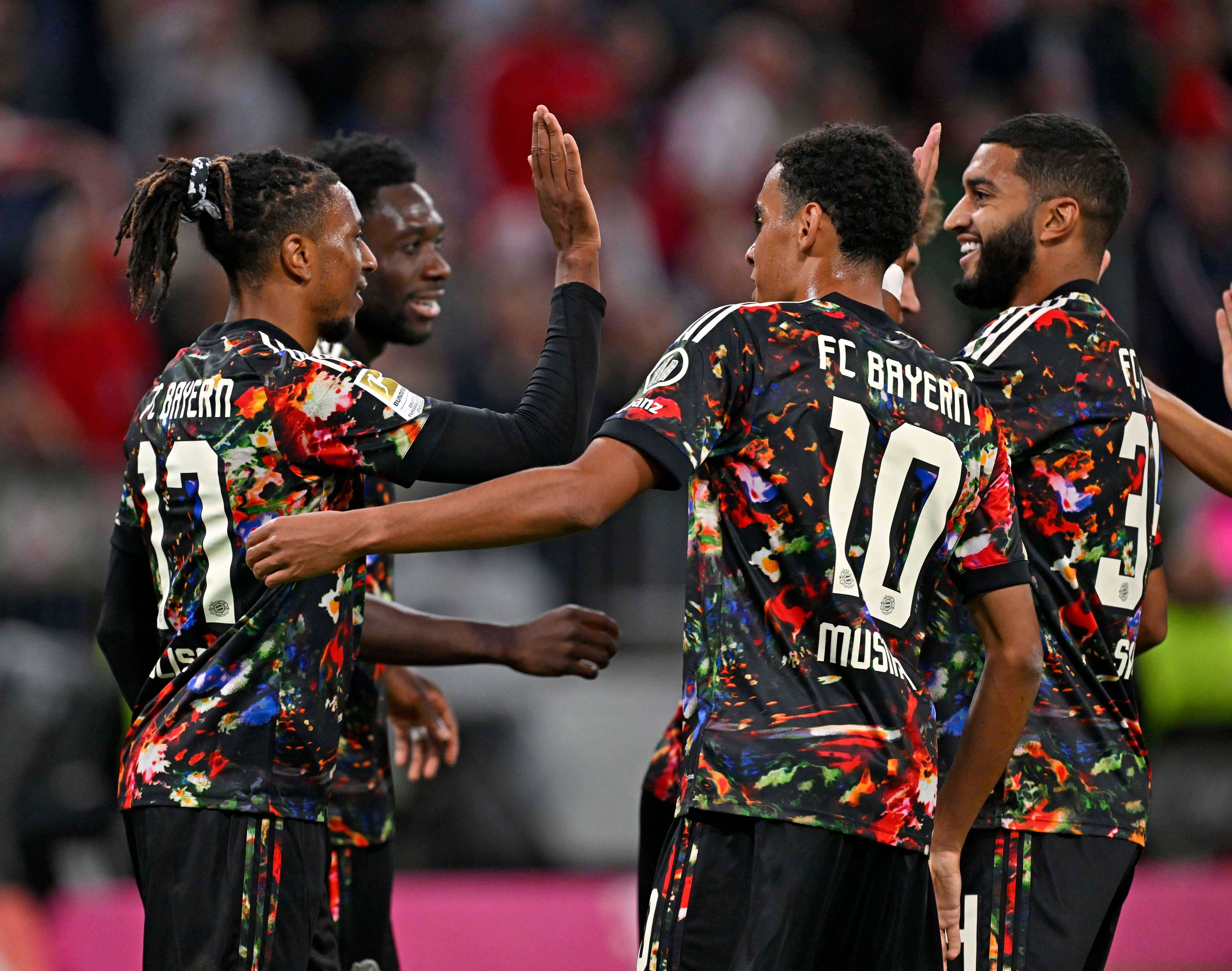 Michael Olise schlägt im bunt gemusterten Wiesn-Trikot des FC Bayern mit Alphonso Davies und Jamal Musiala auf dem Spielfeld ab.