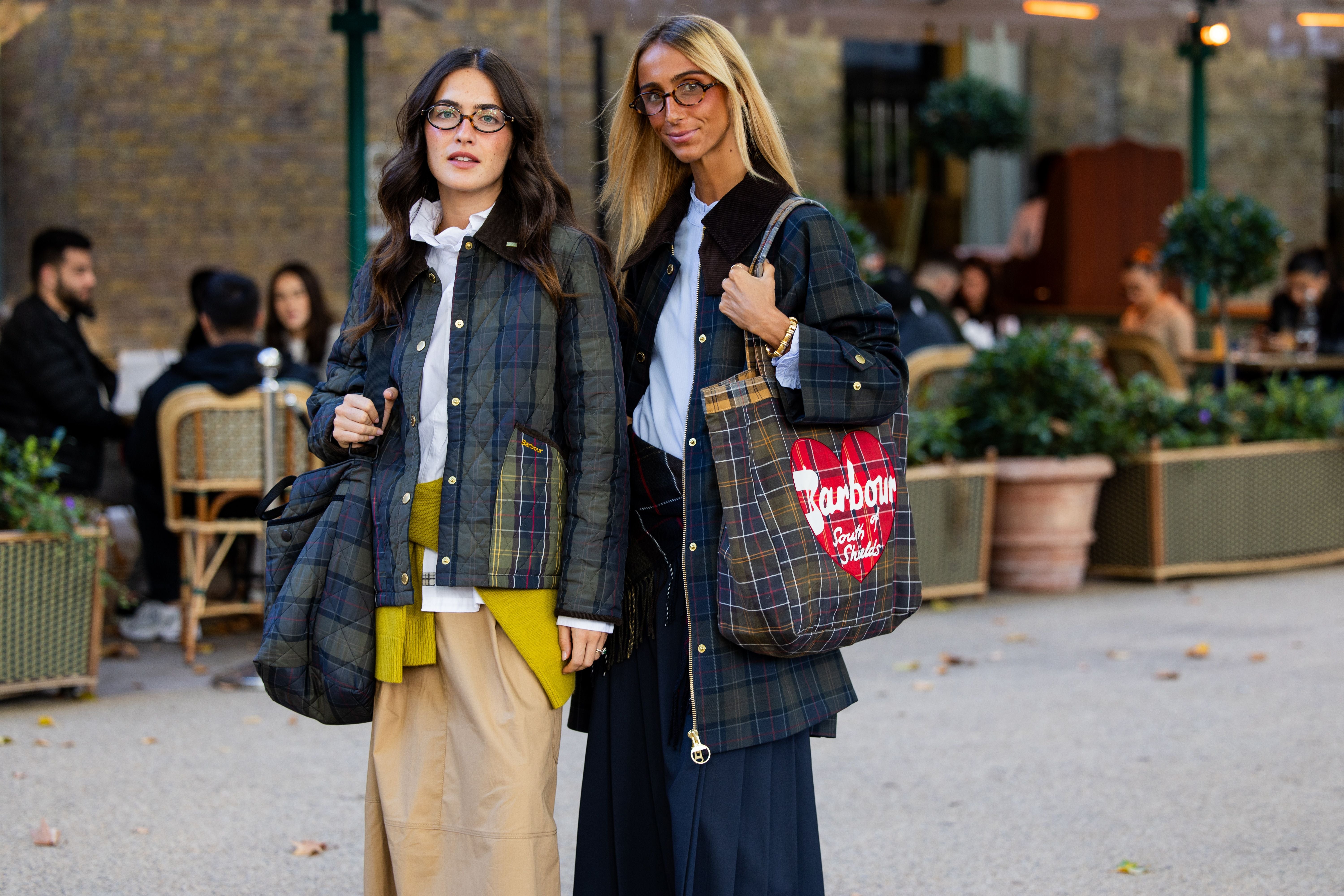 Zwei Frauen in karierten Jacken und Röcken posieren mit Taschen und Brillen auf einer Straße in London.