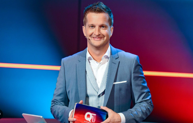 Ein Mann im Anzug steht in einem Fernsehstudio und hält eine Q1-Karte in der Hand.