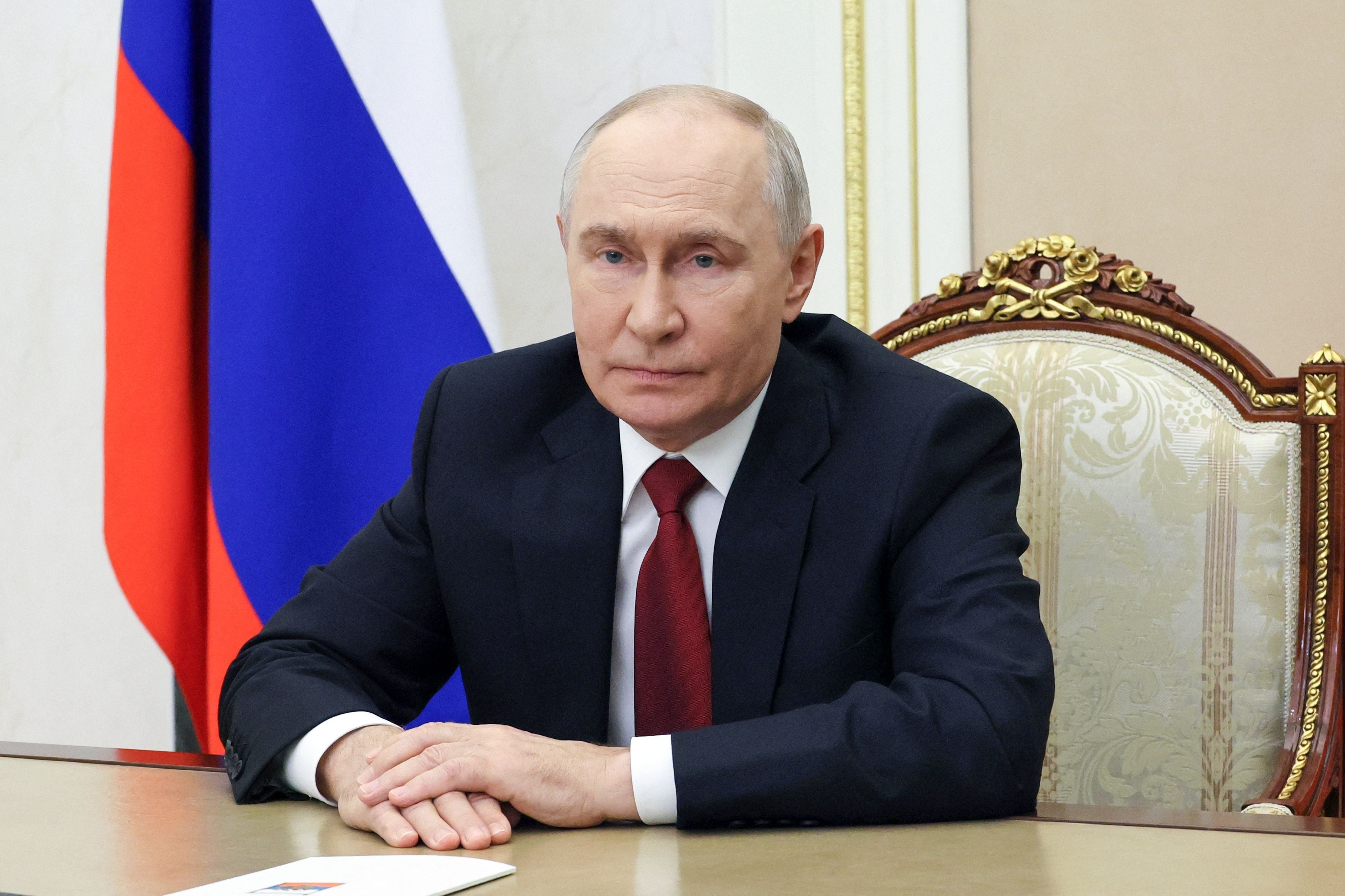 Ein Mann im Anzug sitzt an einem Tisch vor einer russischen Flagge und blickt in die Kamera.