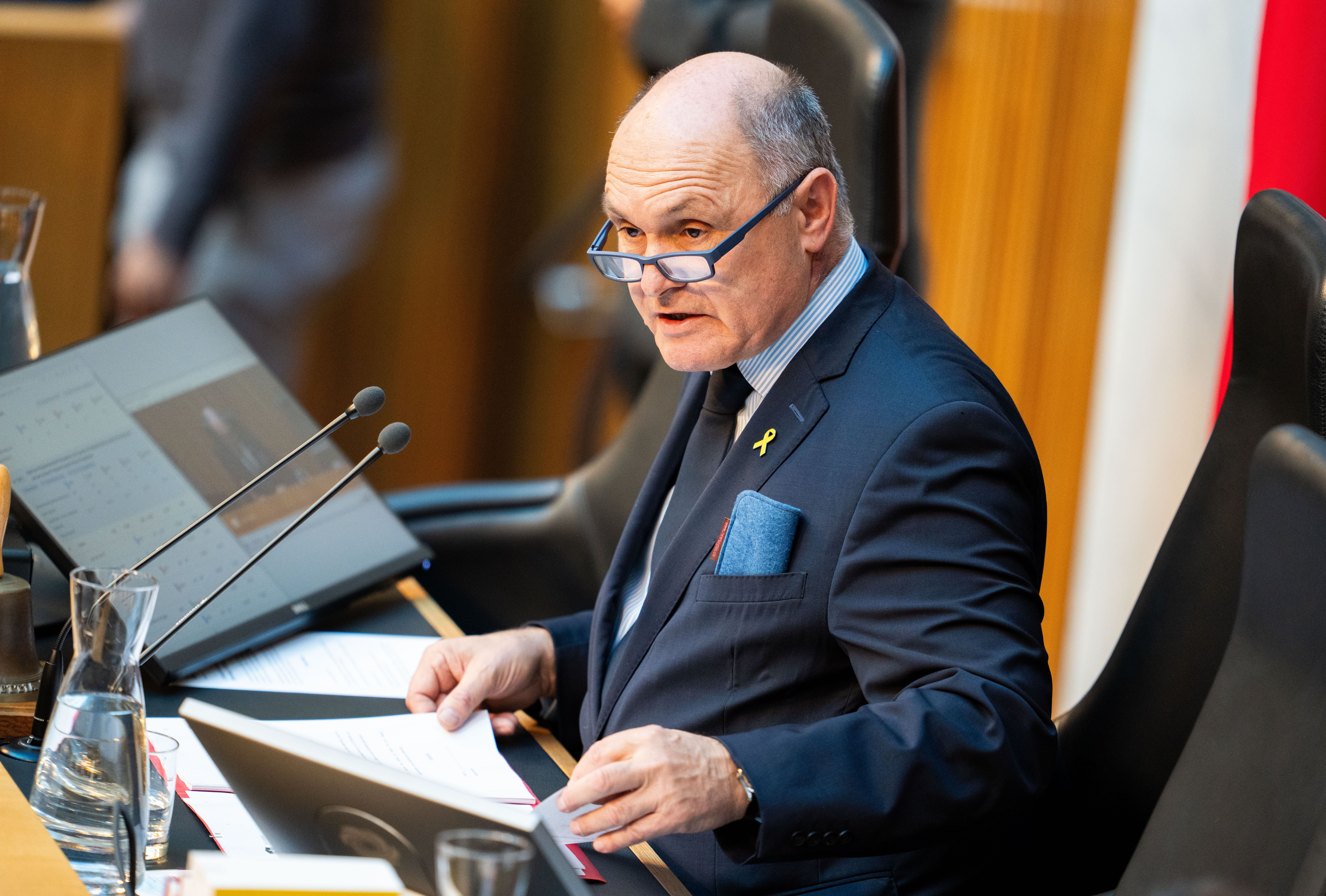 Ein Mann im Anzug sitzt mit Papieren und Brille an einem Schreibtisch im Parlament.