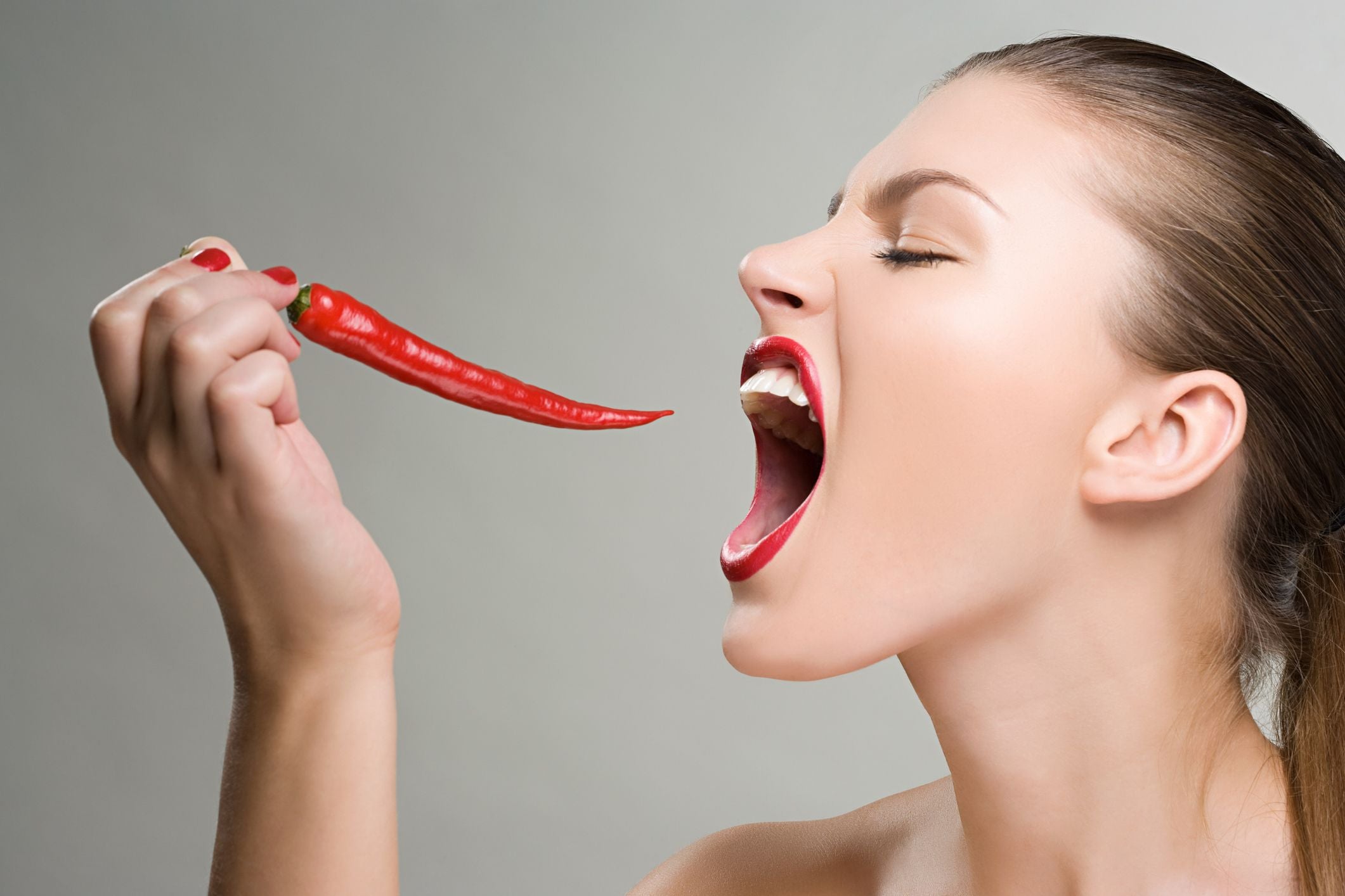 Frau hält eine rote Chilischote und öffnet weit den Mund, als wolle sie hineinbeißen.