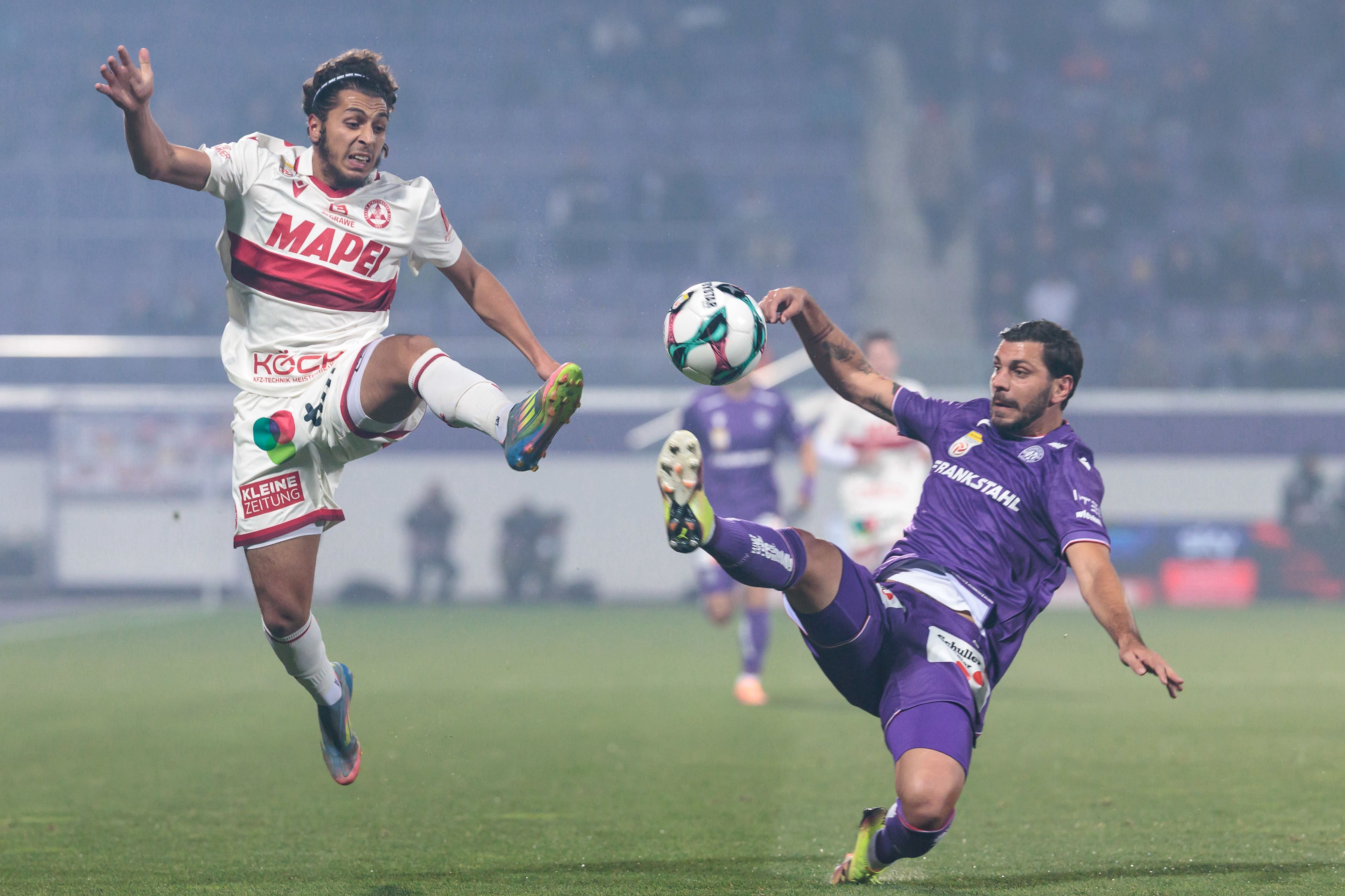 Ein GAK-Spieler im weiß-roten Dress und ein Austria-Wien-Akteur im violetten Trikot kämpfen im Flutlicht mit gestreckten Beinen um einen fliegenden Fußball.