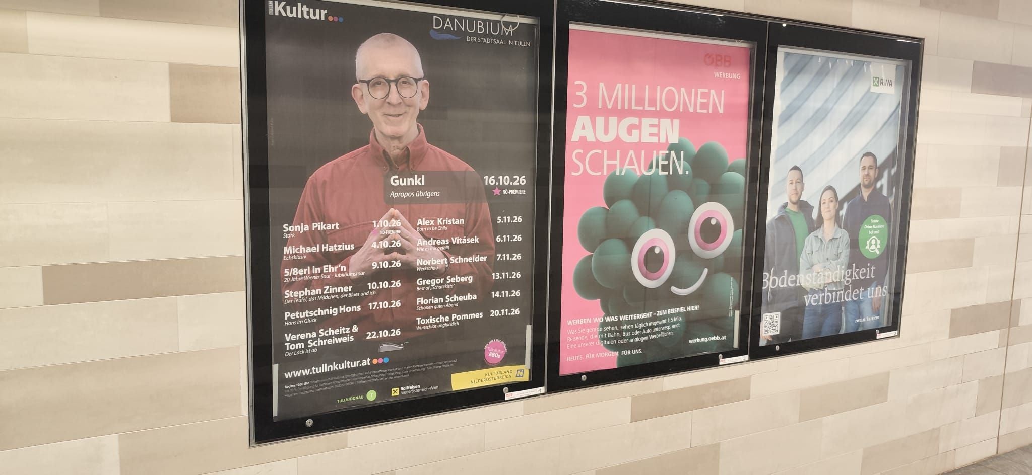 Drei Werbeplakate nebeneinander an einer hellen Fliesenwand in einem öffentlichen Gebäude.