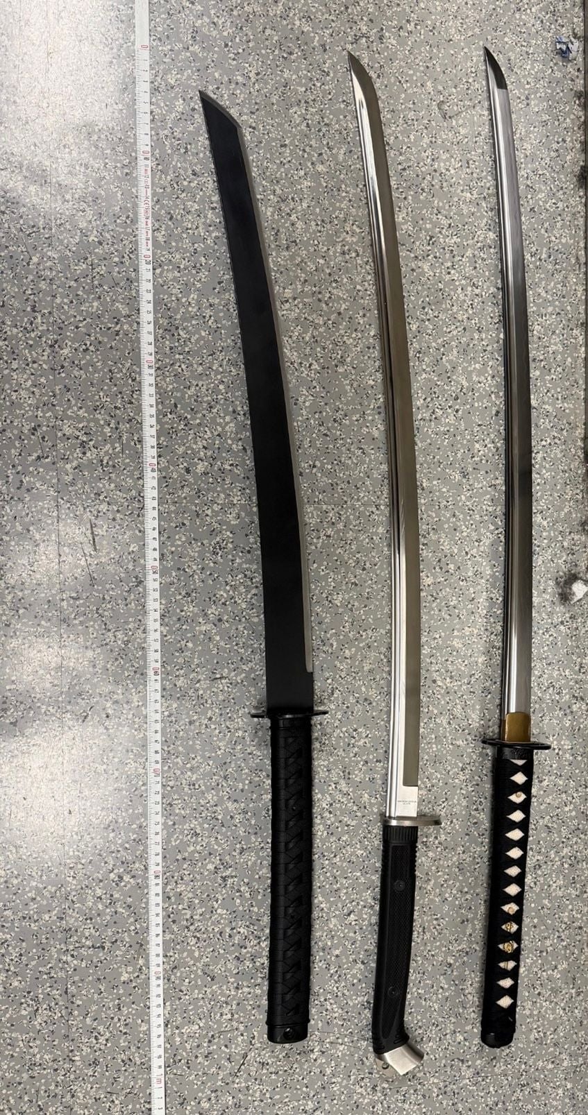 Drei asiatische Schwerter liegen nebeneinander auf einem gefleckten Boden, daneben ein Maßband.