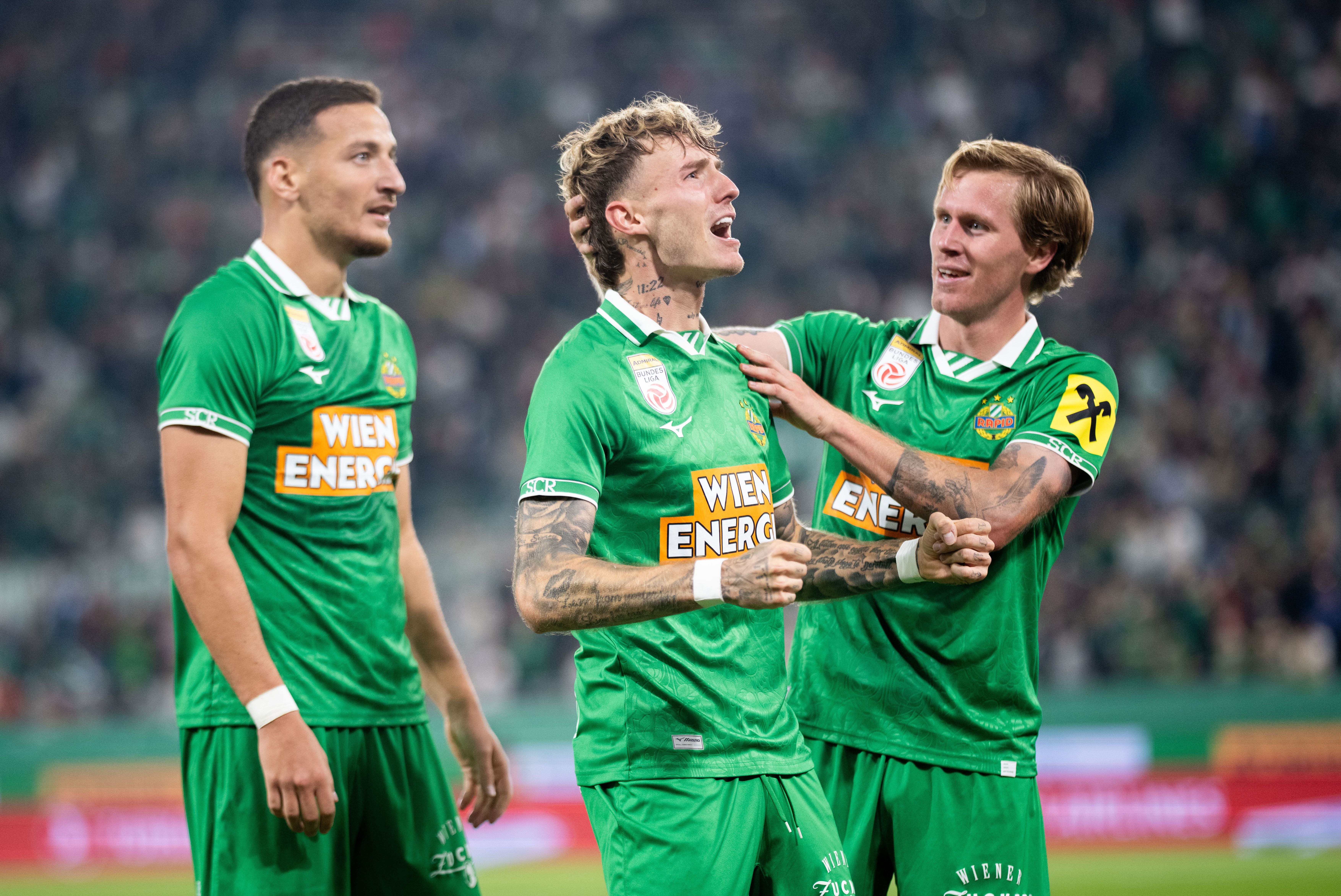 Bendeguz Bolla schreit im grünen Rapid-Trikot seine Freude heraus, während ihn Tonni Adamsen am Kopf umarmt und Ercan Kara daneben steht.