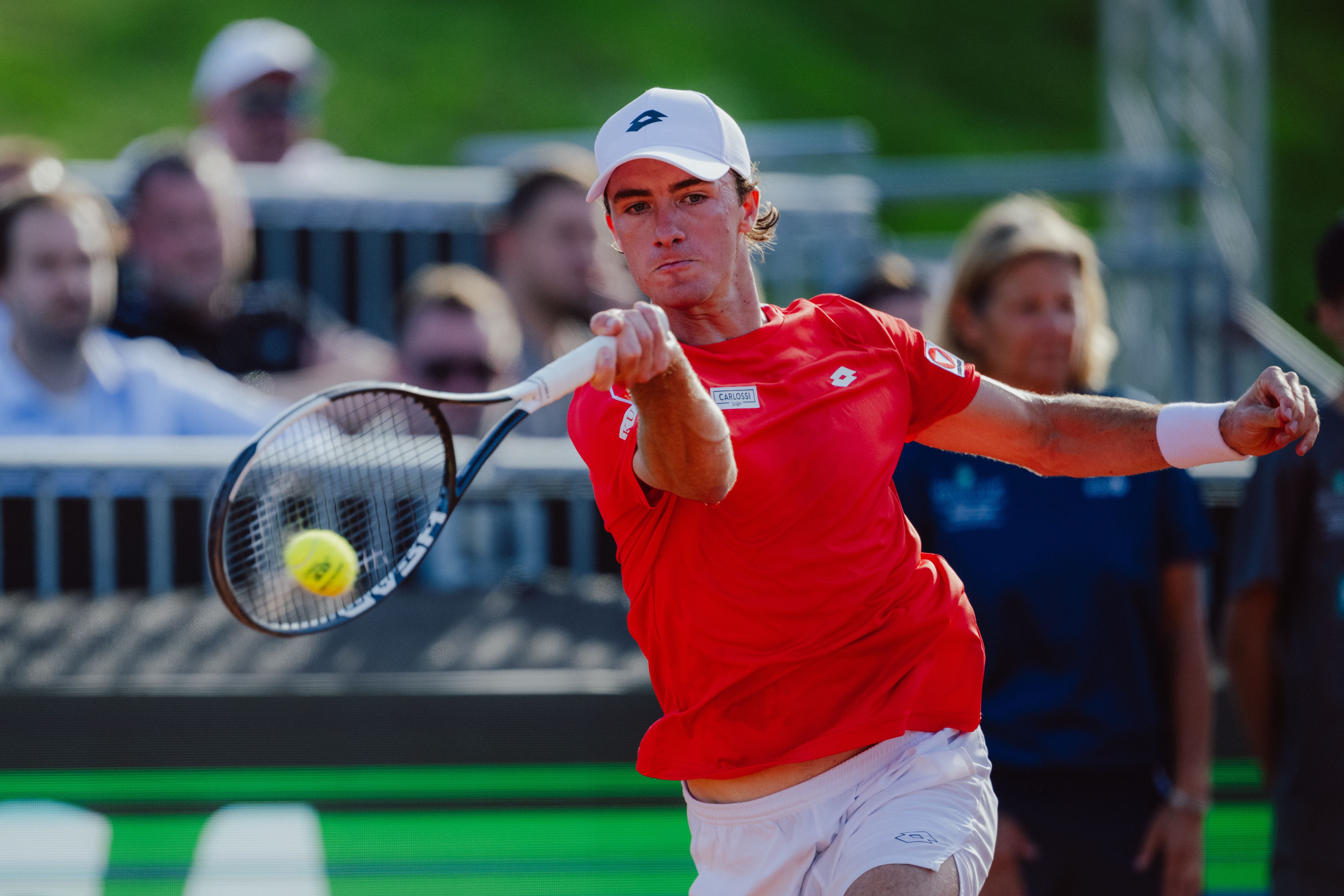Tennisspieler in rotem Shirt schlägt den Ball während eines Matches, Zuschauer im Hintergrund.