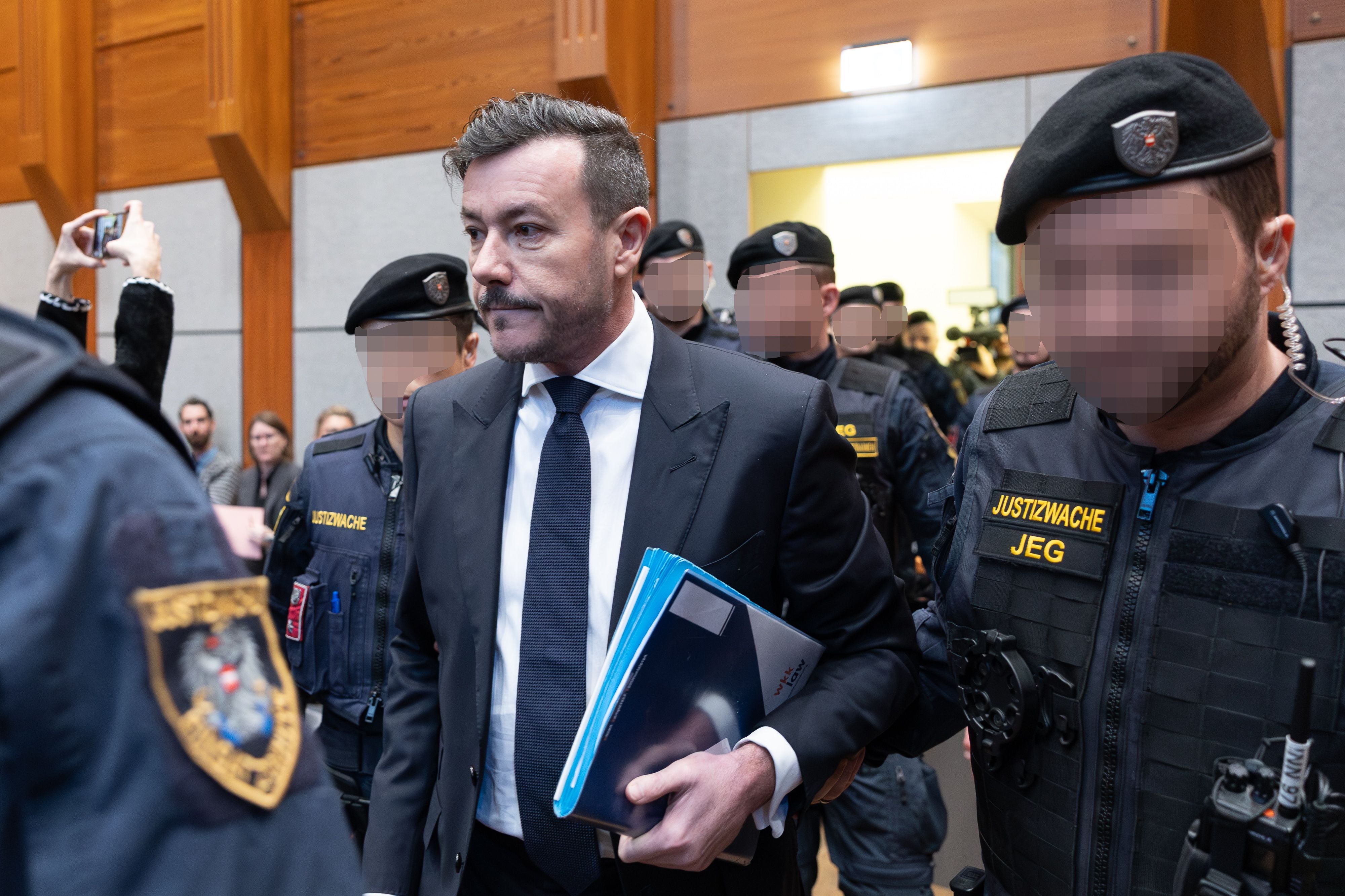 Rene Benko wird von Justizwachebeamten bei Prozessbeginn im Gerichtssaal begleitet.
